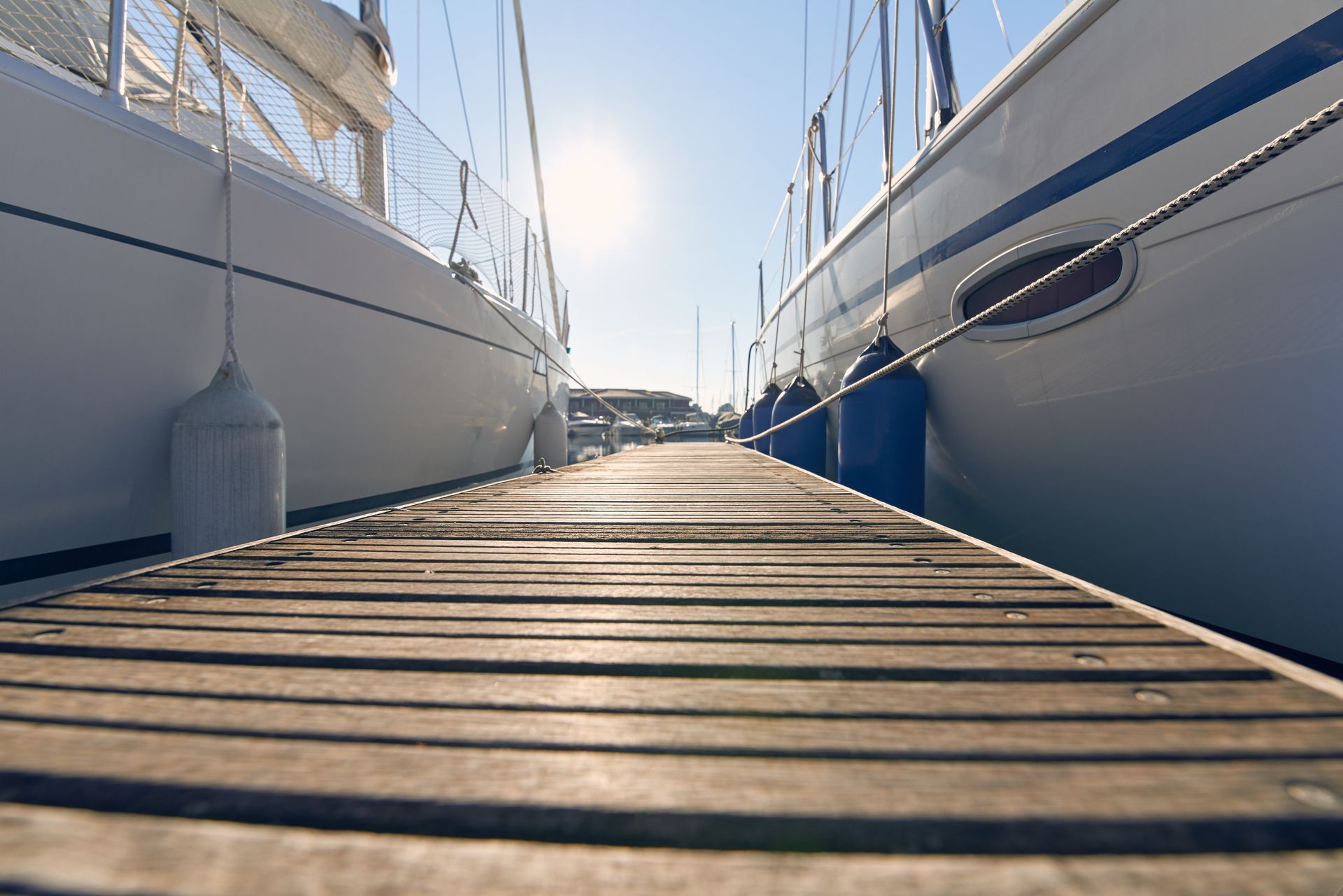 Wooden dock between two white sailboats at a marina, with the sun shining ahead.