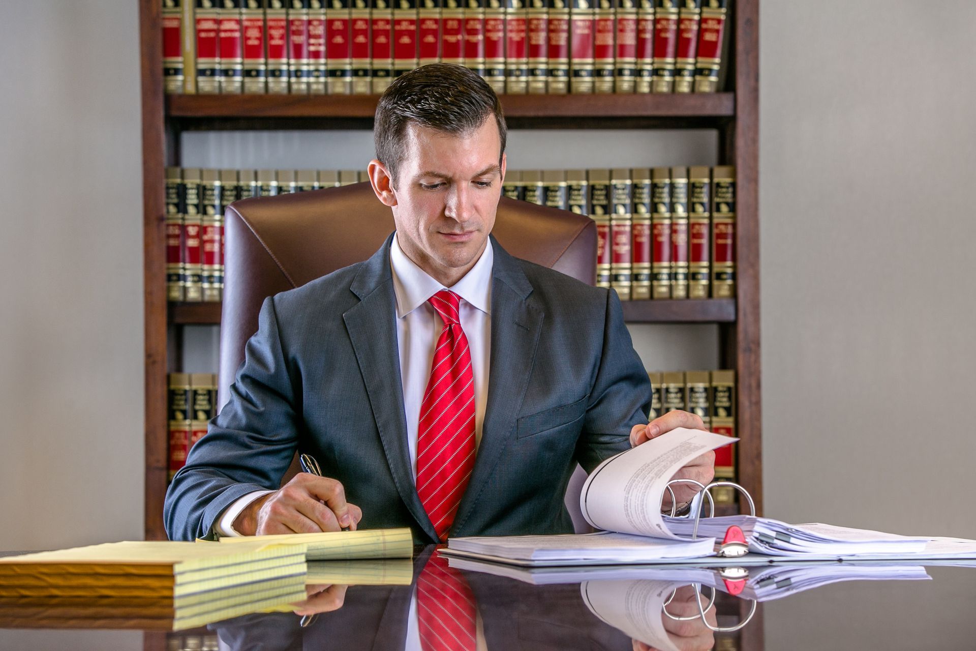 A man in a suit and tie is sitting at a desk in front of a bookshelf.