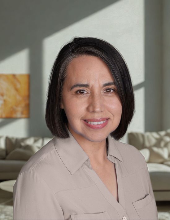 A woman with tan shirt is standing in a house in front of a couch with a painting on the wall.