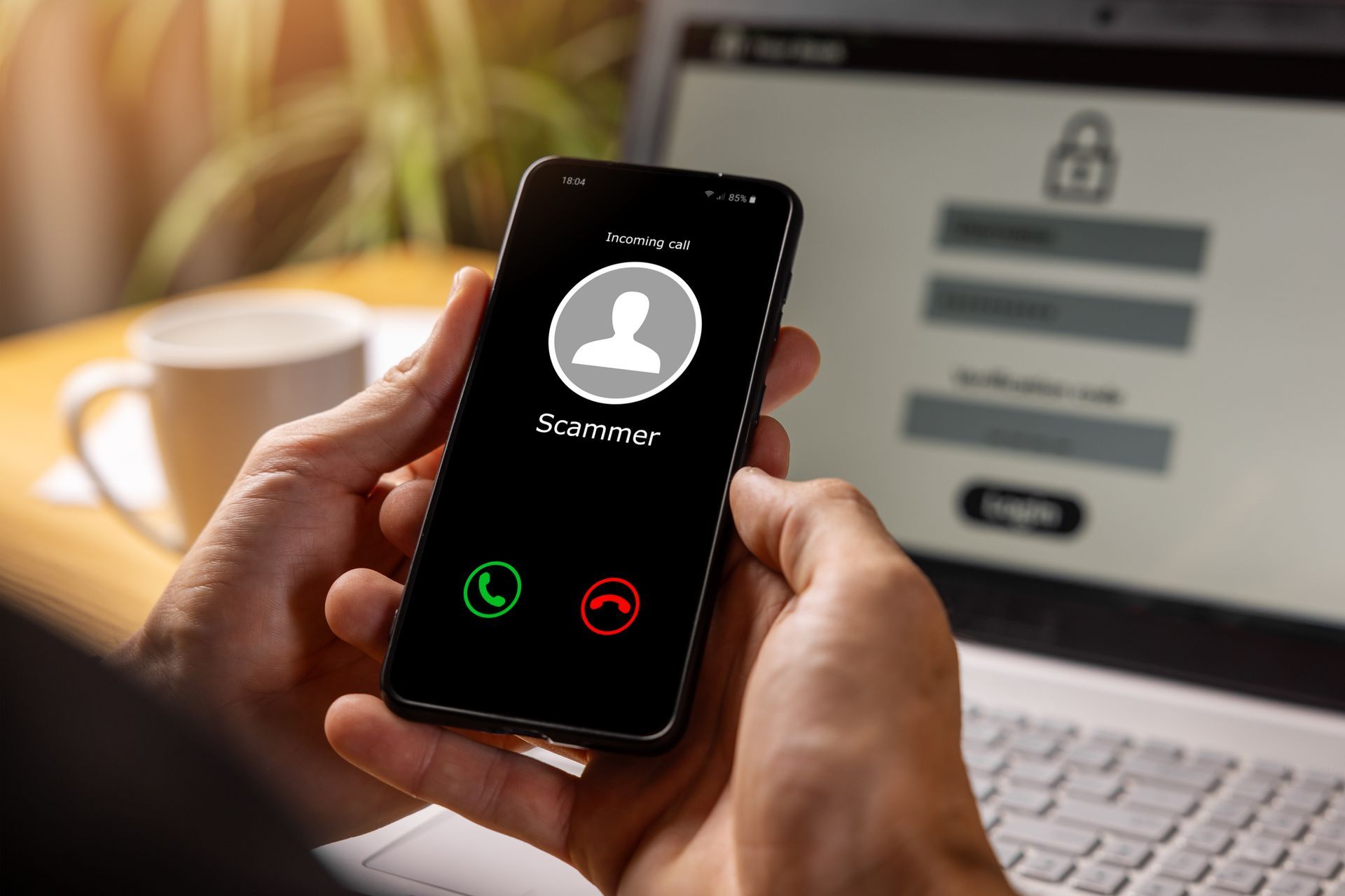 Hand holding a smartphone with an incoming call screen in front of a laptop login page