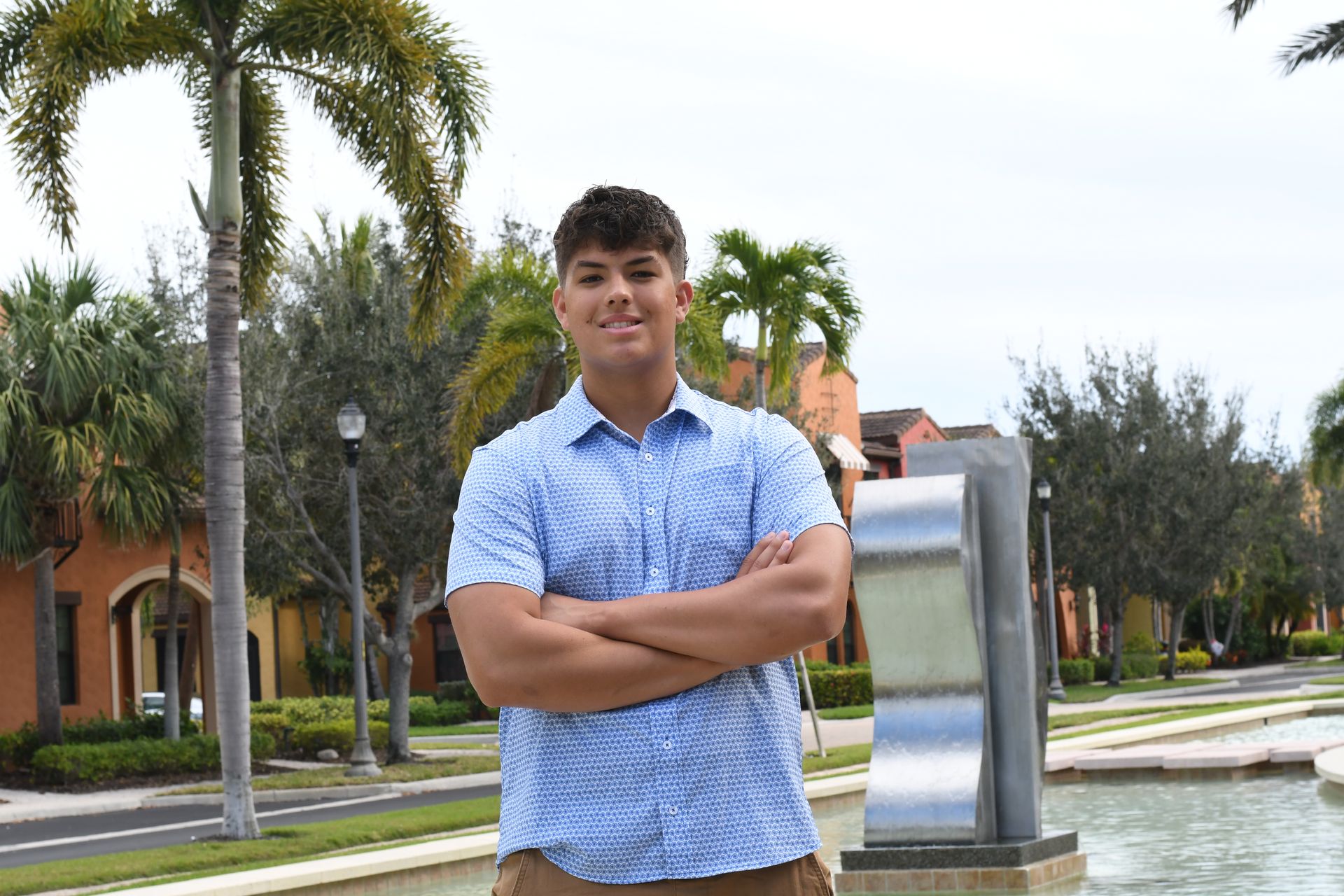 Person standing with arms crossed near a fountain and palm trees outdoors