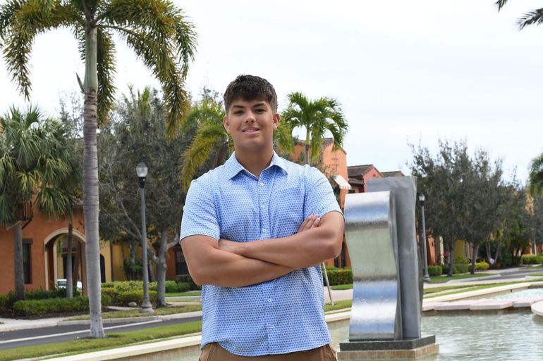 Person standing with arms crossed near a fountain and palm trees outdoors