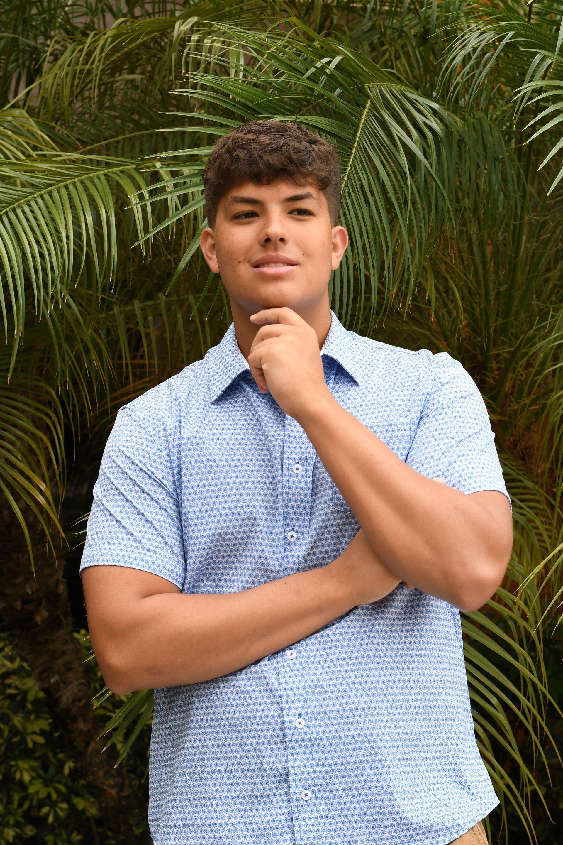 Person in a blue patterned shirt posing outdoors in front of palm leaves.