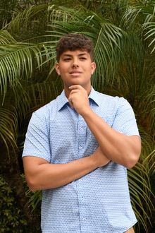Person in a blue patterned shirt posing outdoors in front of palm leaves.