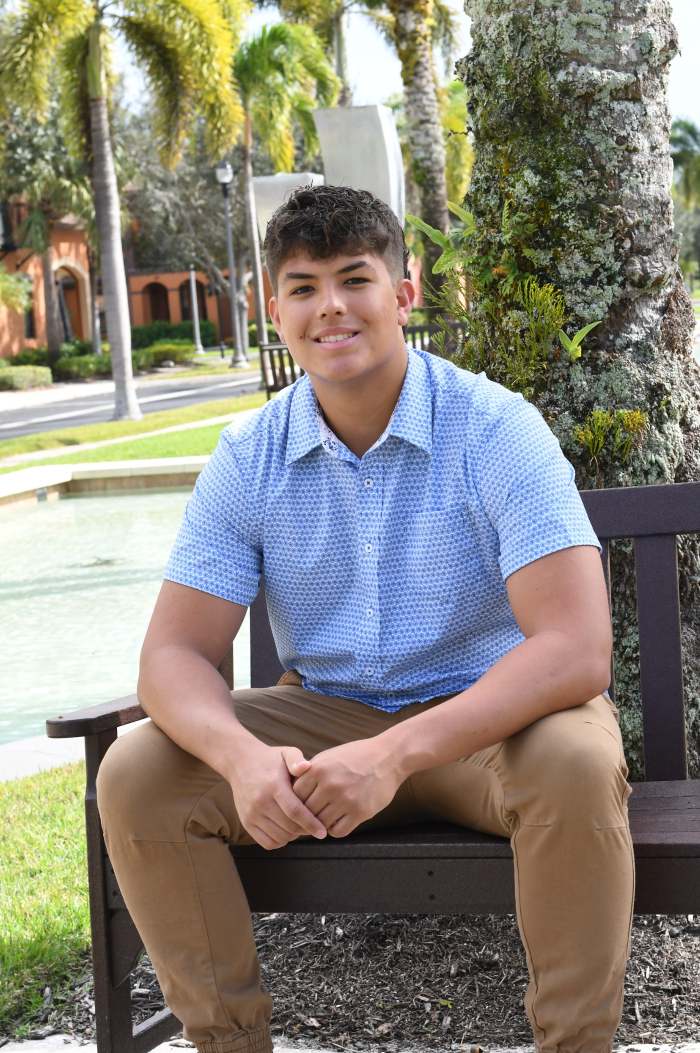 Person in a blue patterned shirt and tan pants sitting on a bench outdoors in a park.