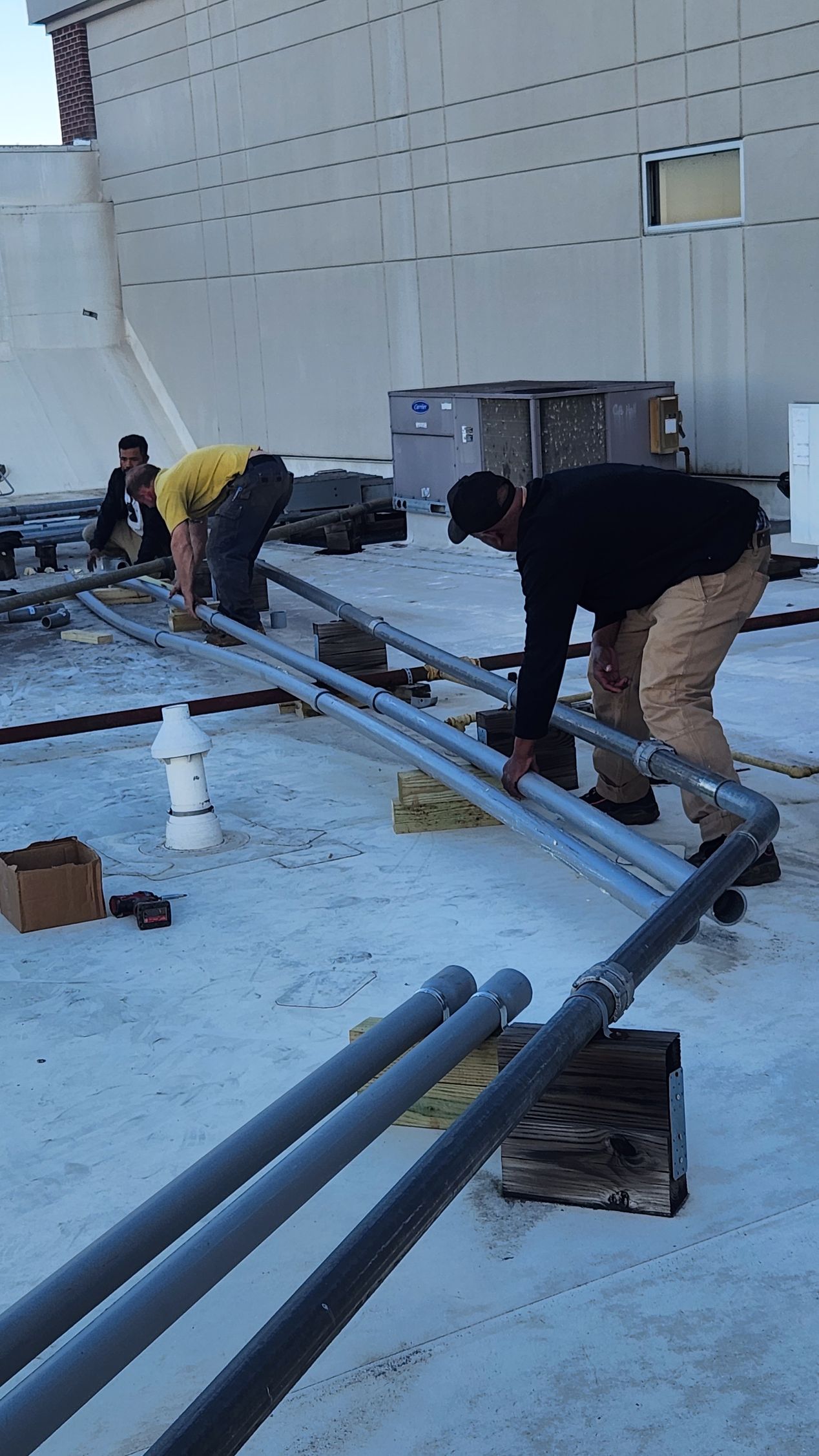 Workers installing metal pipes on a flat rooftop, near a building. They are wearing work clothes.