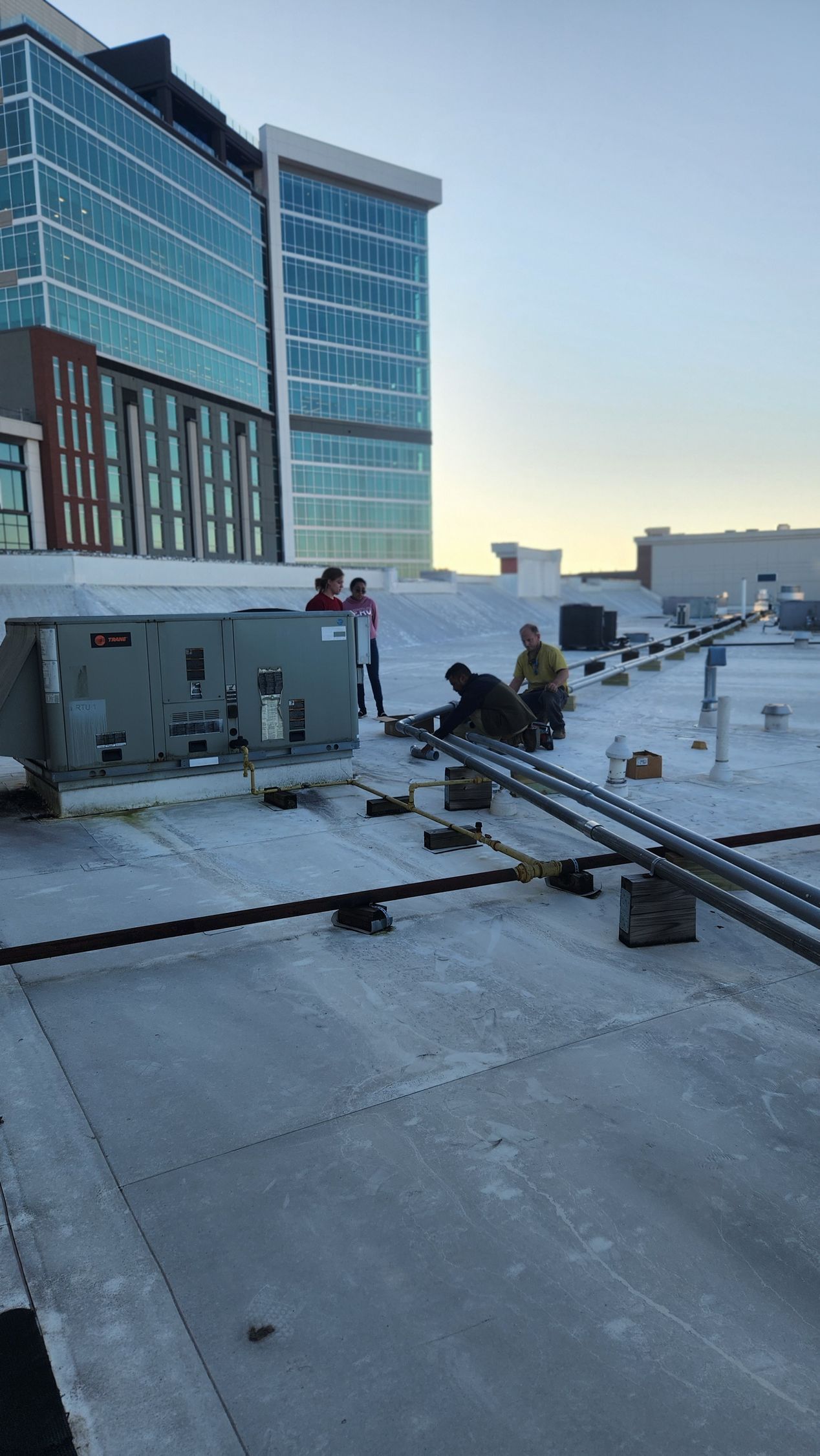 Workers on a rooftop install equipment near a large building. The sky is clear, daytime.
