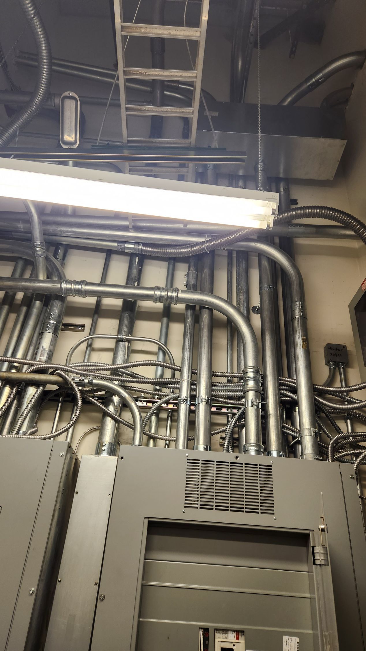 Electrical conduits and wiring on a wall, with a metal ladder and overhead lighting visible.