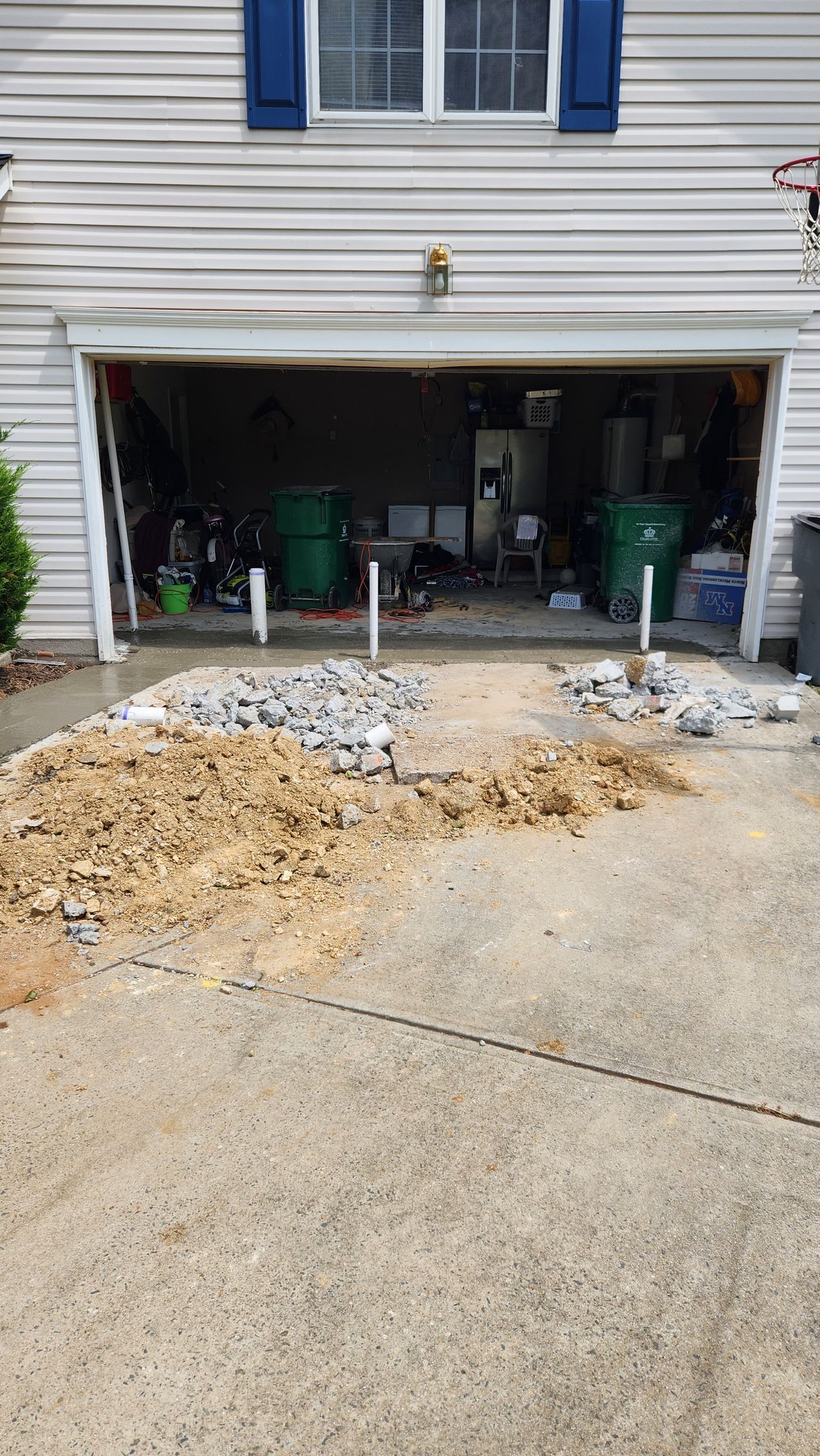 Driveway construction: Excavated gravel and dirt in front of a garage, with two green trash bins visible inside.