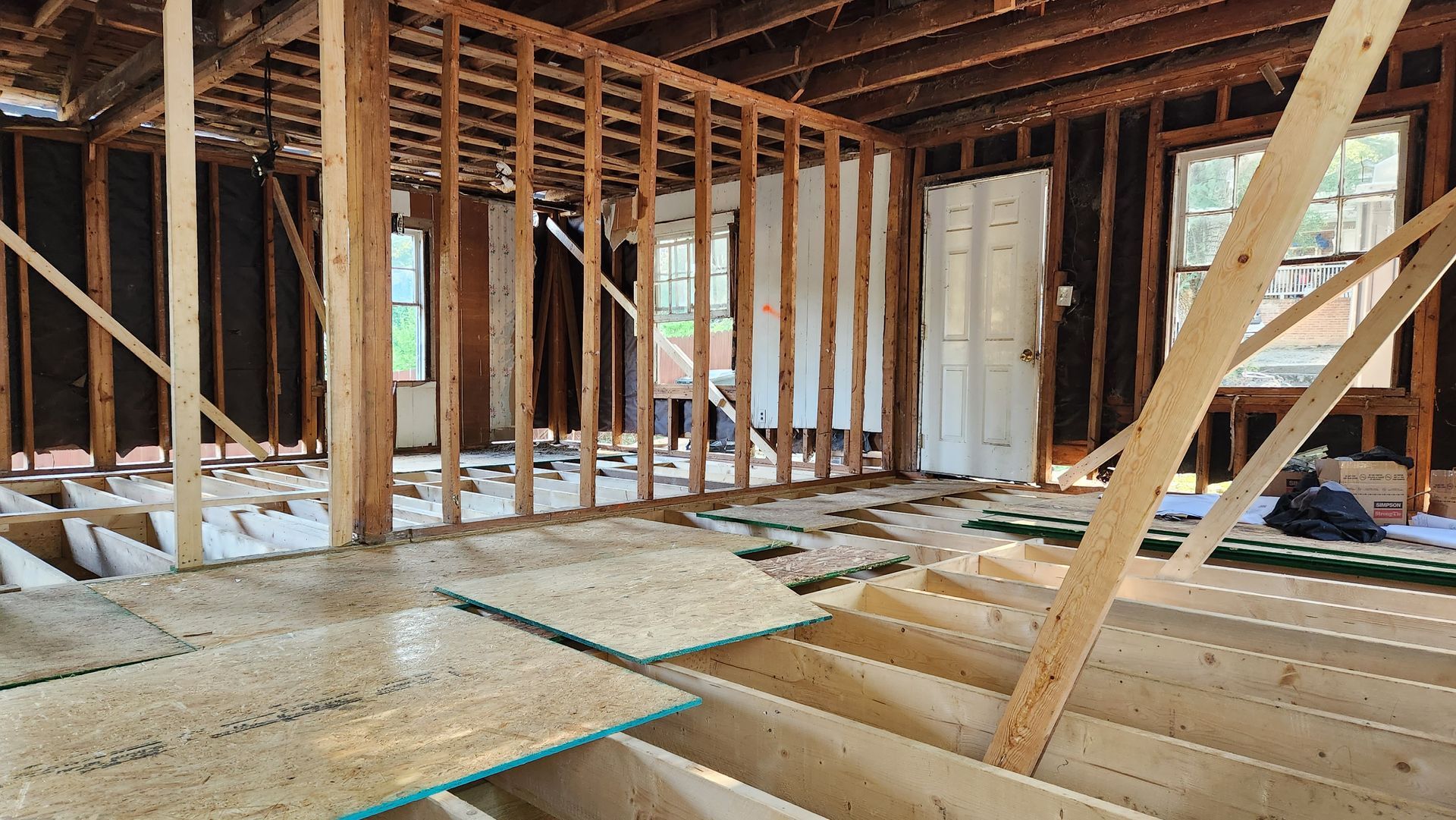 Interior framing of a room under construction with exposed wooden studs, floor joists, and partially installed subflooring.