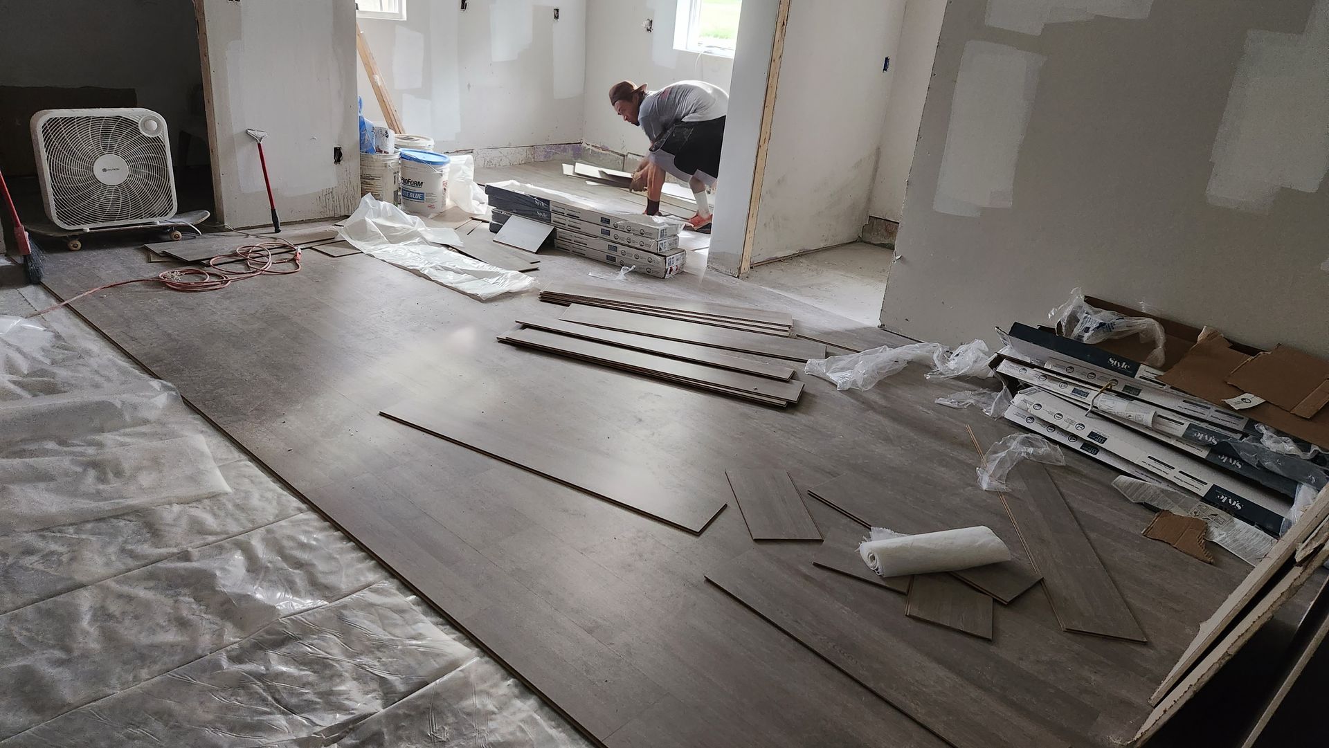 Person installing gray flooring in a room with a partially tiled wall; tools and materials scattered.