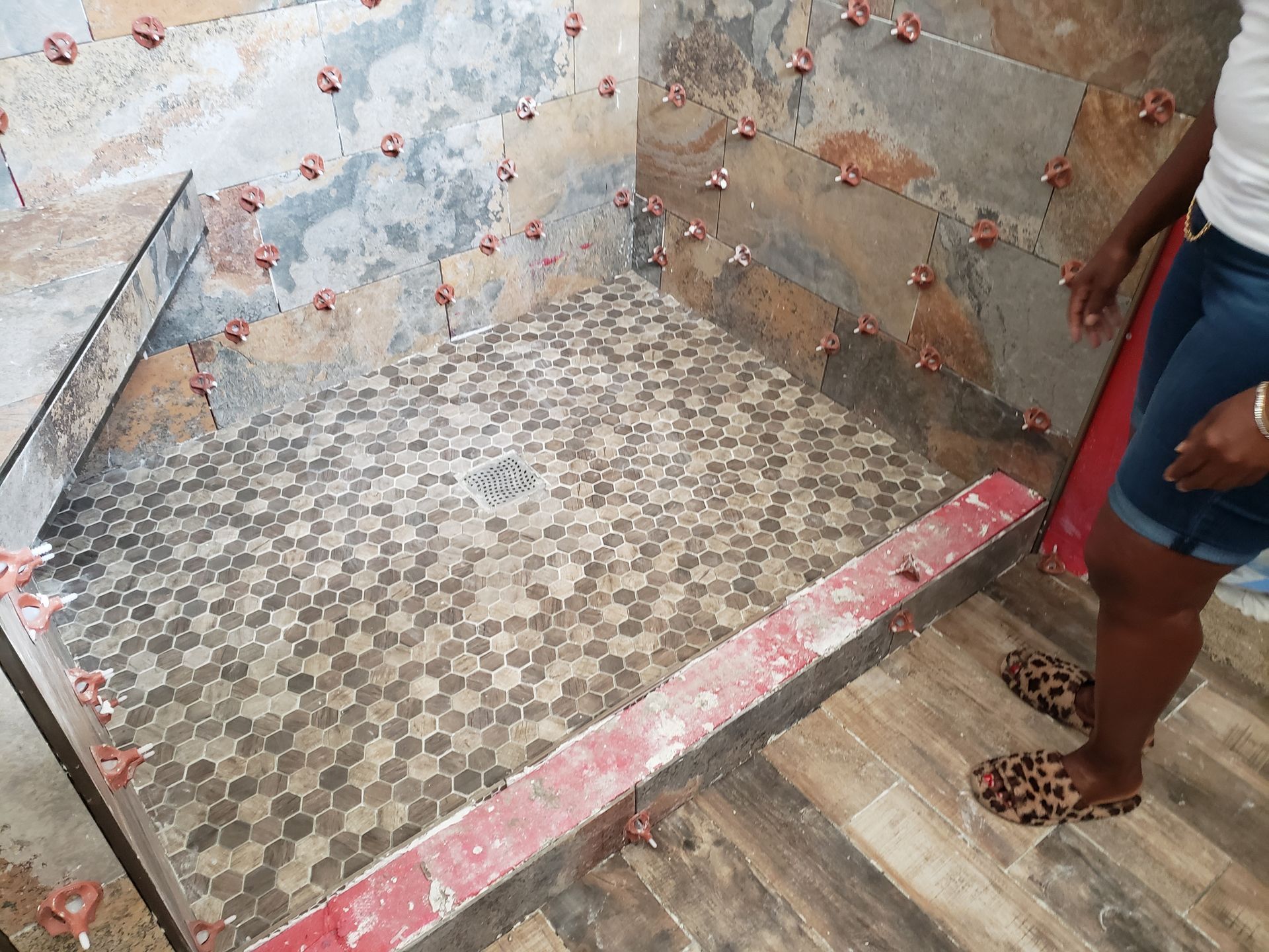 Shower in progress with hexagon tiles on the floor and slate-like tiles on walls; person standing nearby.