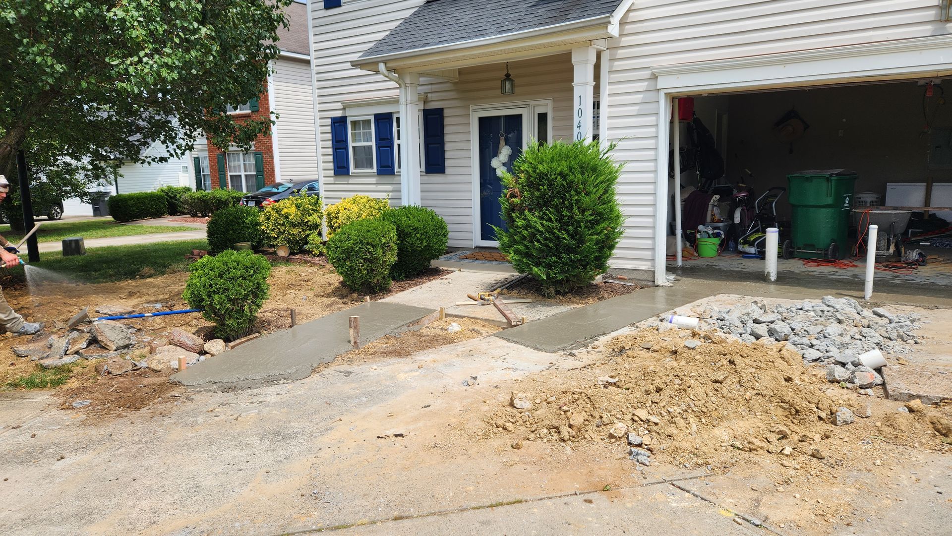 Concrete driveway being constructed in front of a house, with bushes and dirt piles.