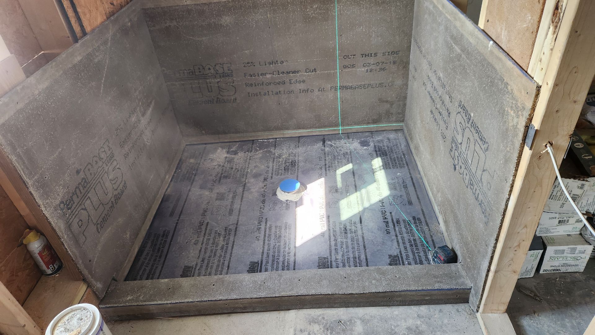 Shower stall construction: gray walls, black floor, blue drain, sunlight, and wooden framing.
