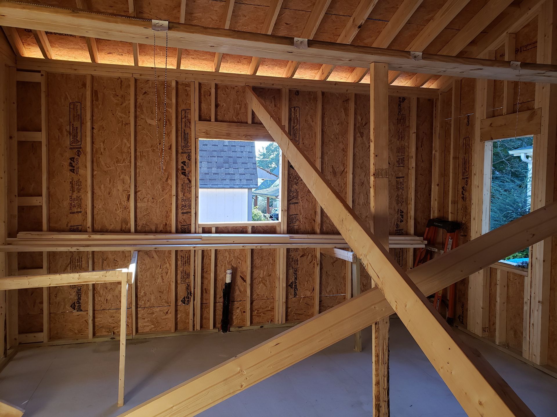 Interior of a building under construction, wooden frame, two windows, rafters, OSB sheathing, unfinished concrete floor.
