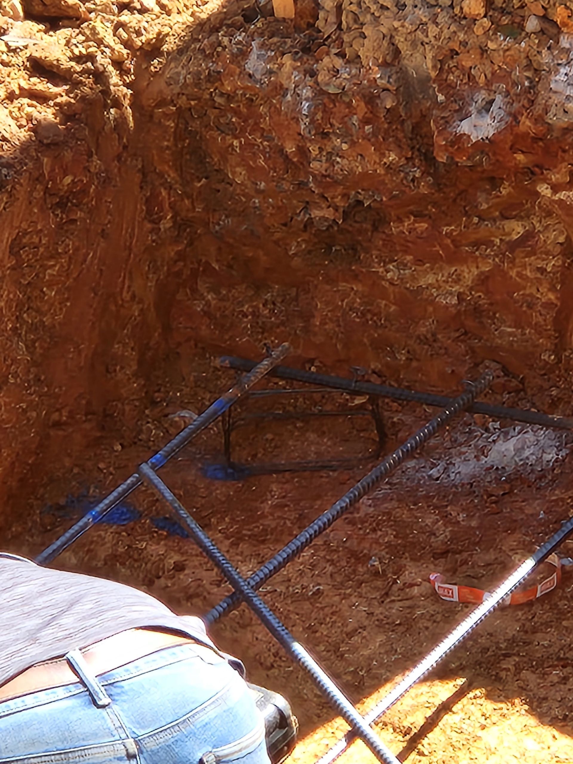 Person in jeans working on rebar frame in an excavated pit with brown soil.