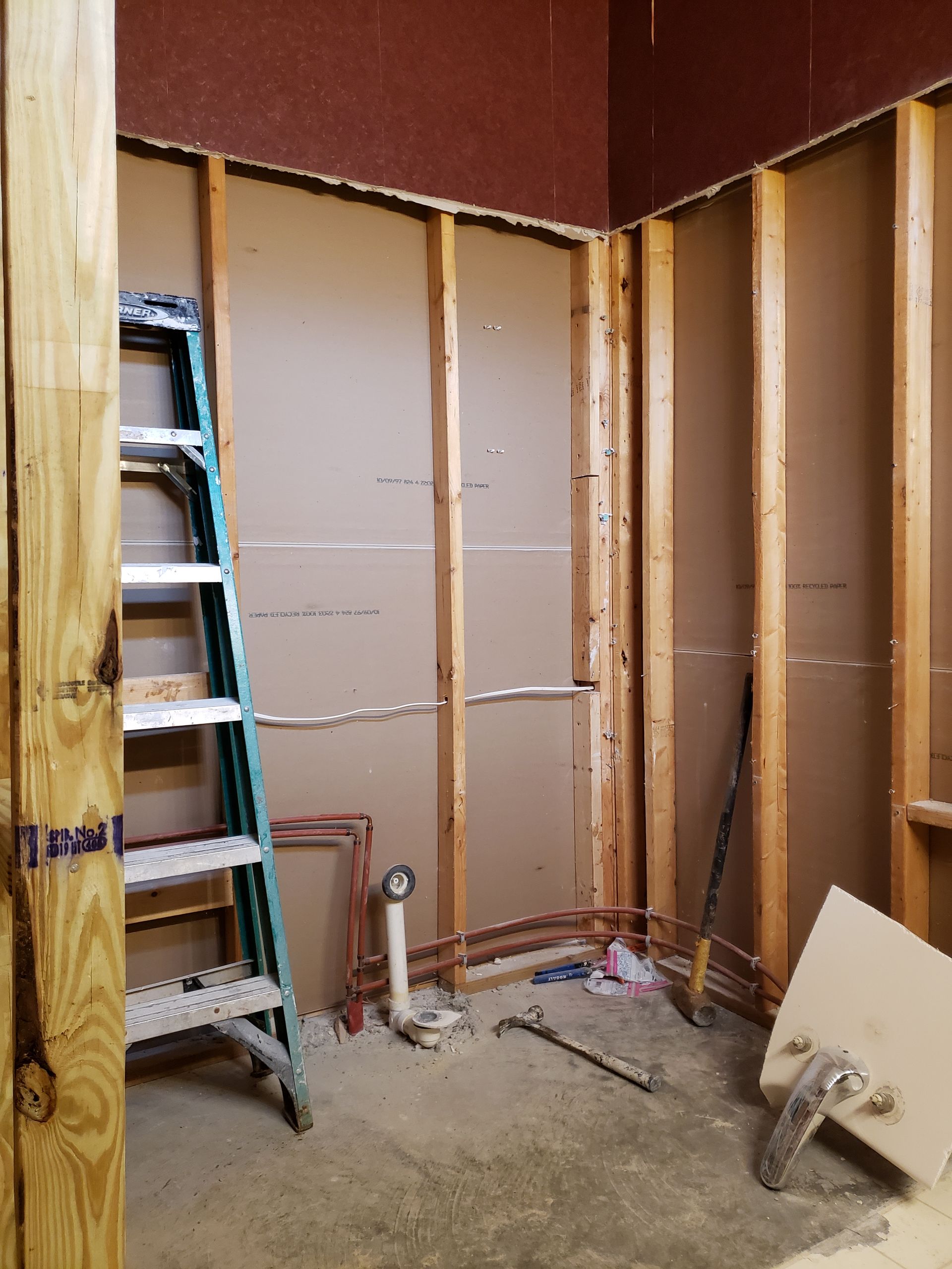 Bathroom under construction: exposed studs, copper pipes, ladder, drywall, and tools.