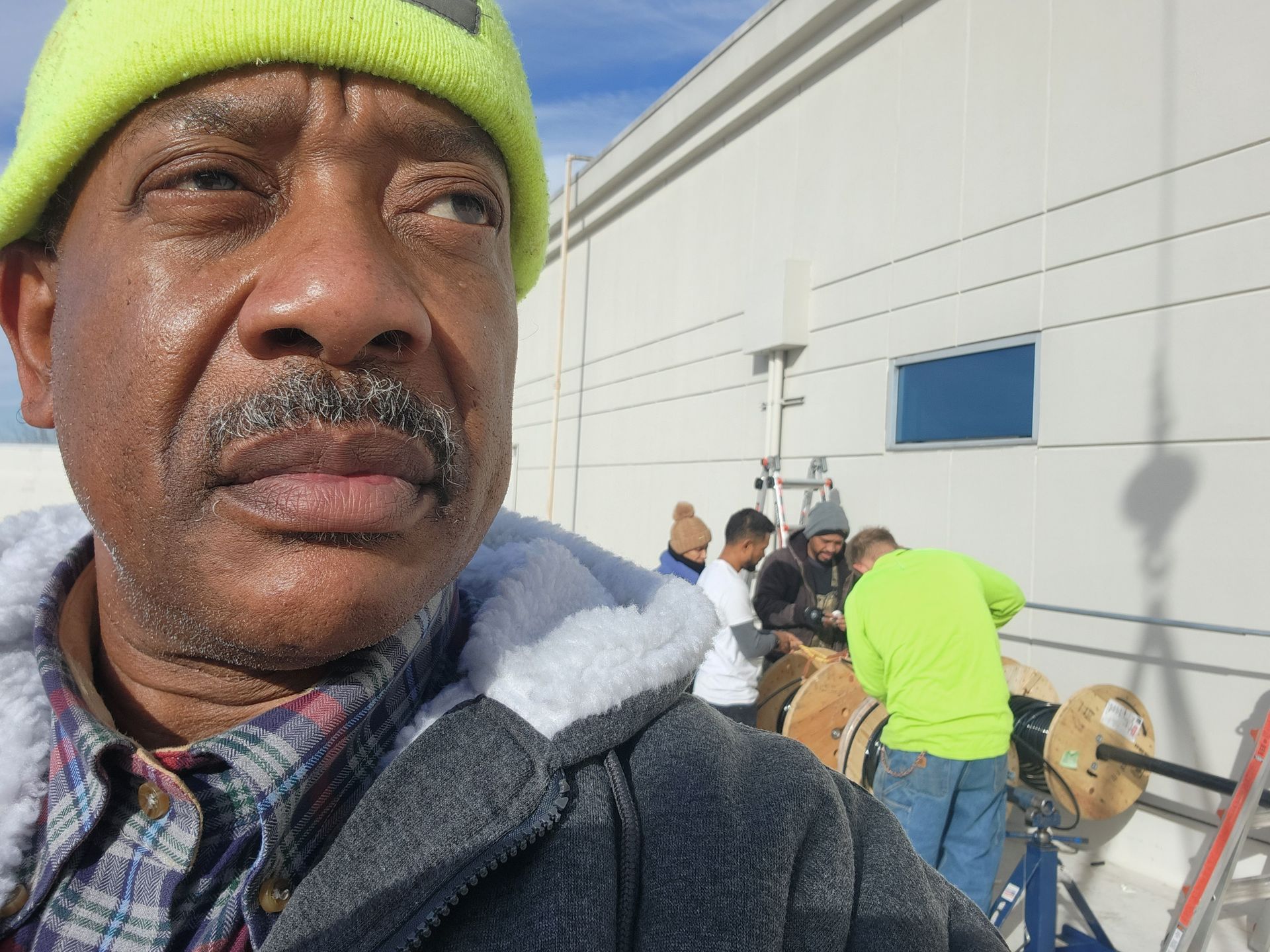Man in a yellow hat looks toward the side as workers spool cable on a rooftop.