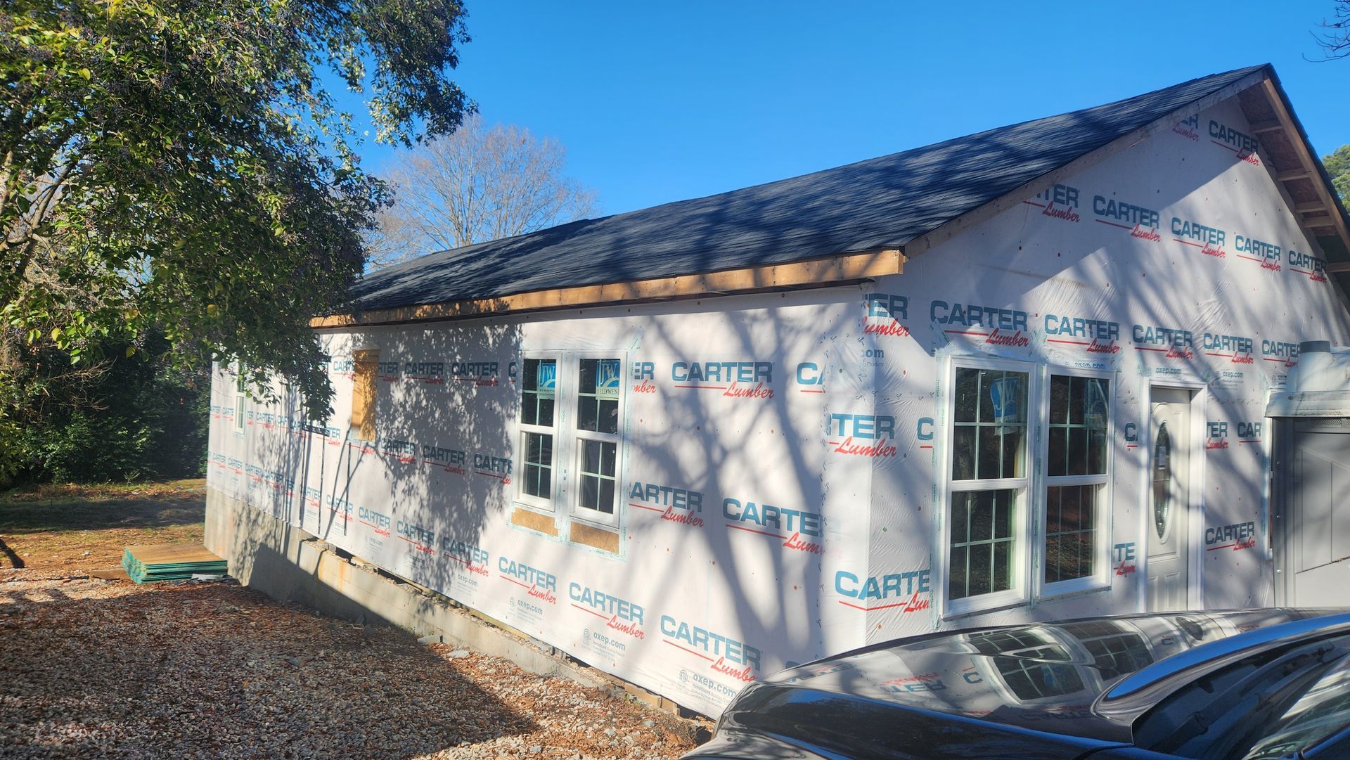 Building under construction with dark roof, windows, and white house wrap on a sunny day.