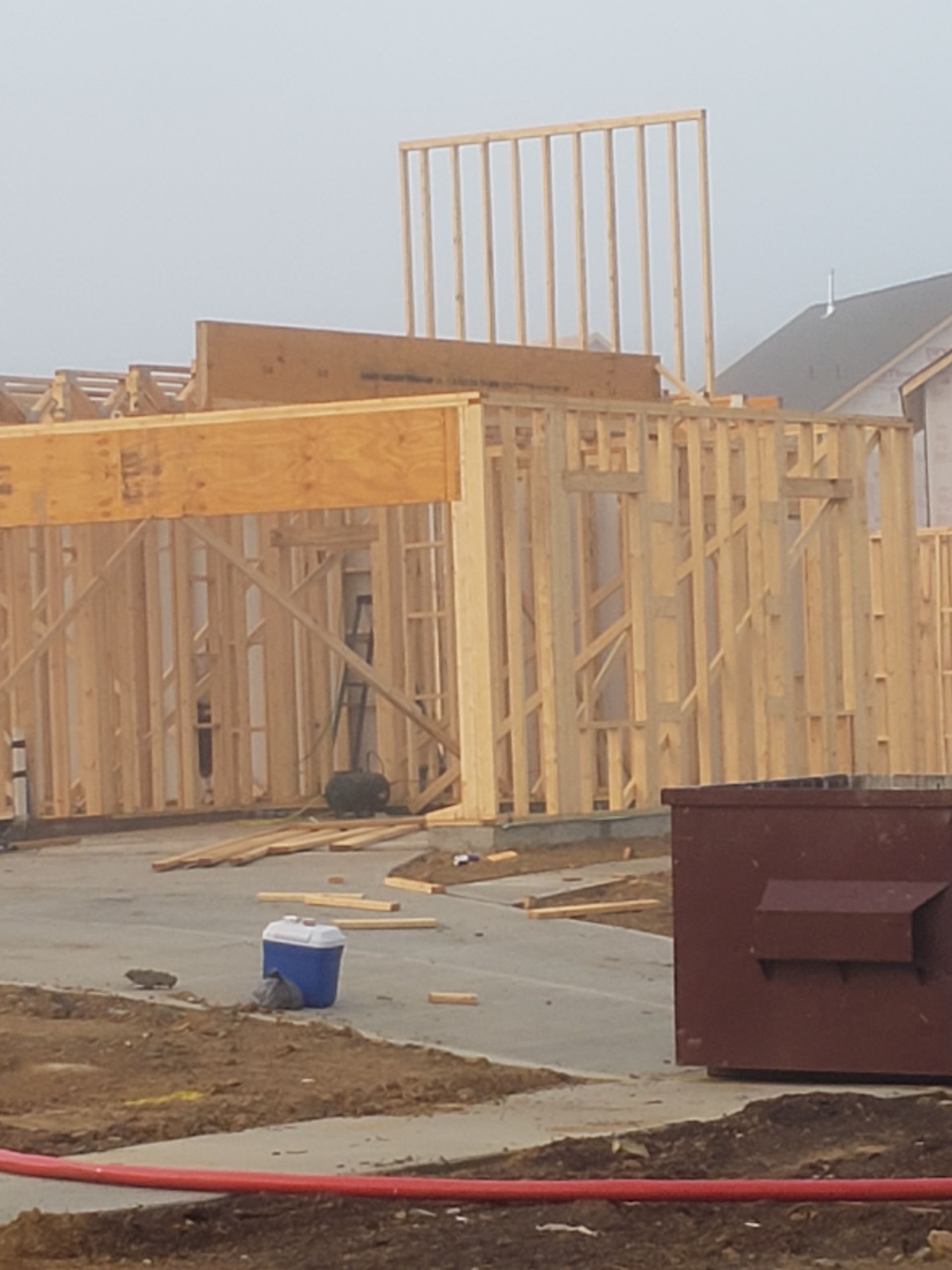 Framing of a new house under construction on a cloudy day, with a dumpster and debris in the foreground.