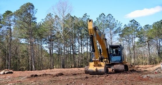 A yellow excavator is moving dirt in a field with trees in the background.
