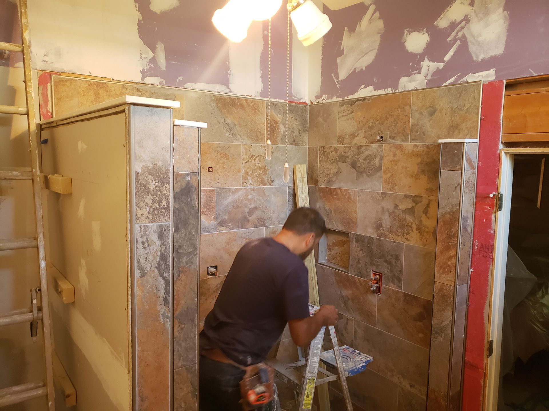 Man tiling shower walls, using a ladder. Bathroom under construction, stone tiles, neutral tones.