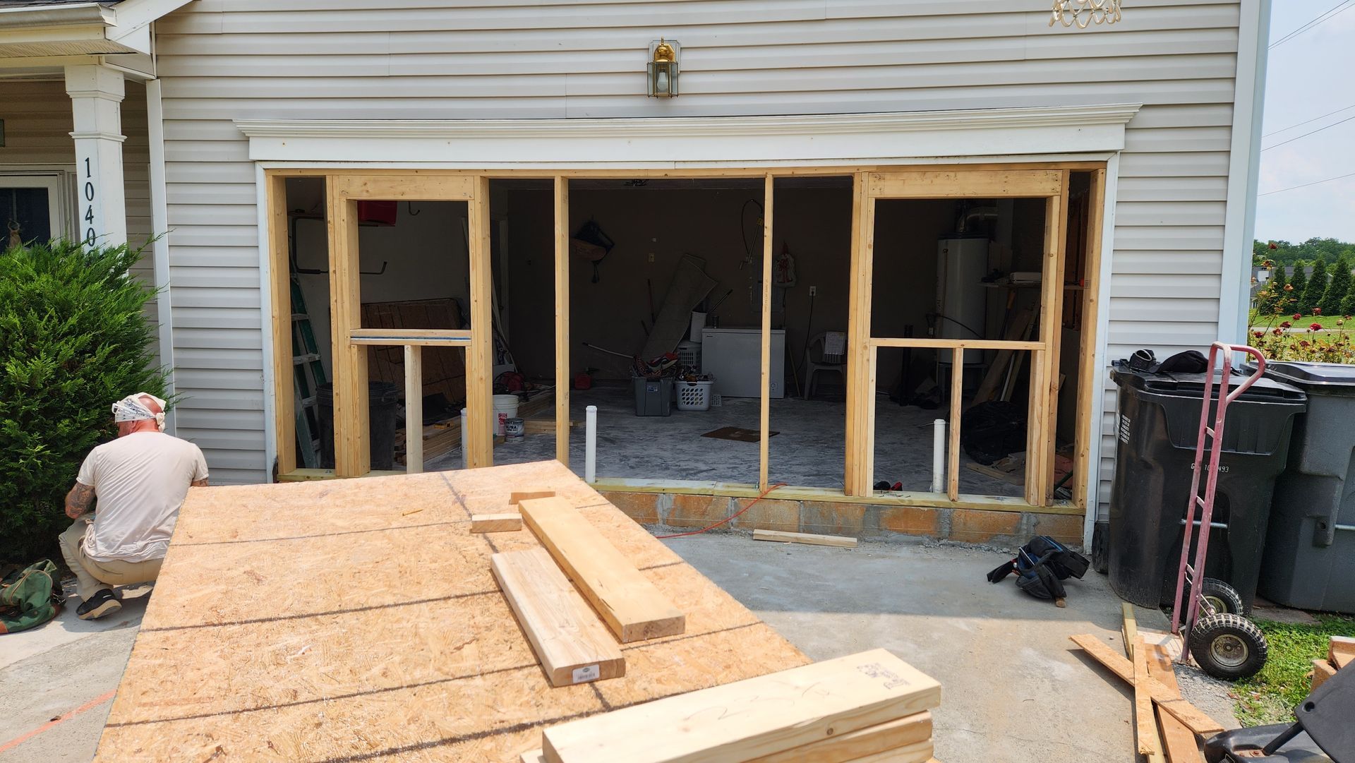 Man working on garage door frame; wood, tools, and construction in progress outside.