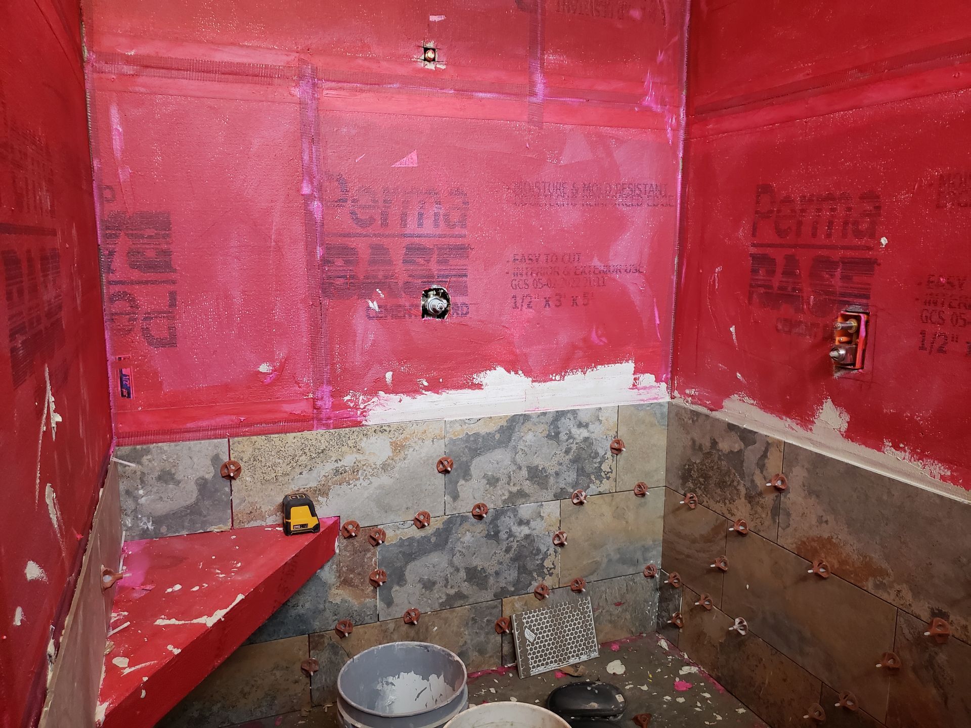 Bathroom shower under construction; red waterproof membrane applied to walls, exposed cement floor with tile spacers.