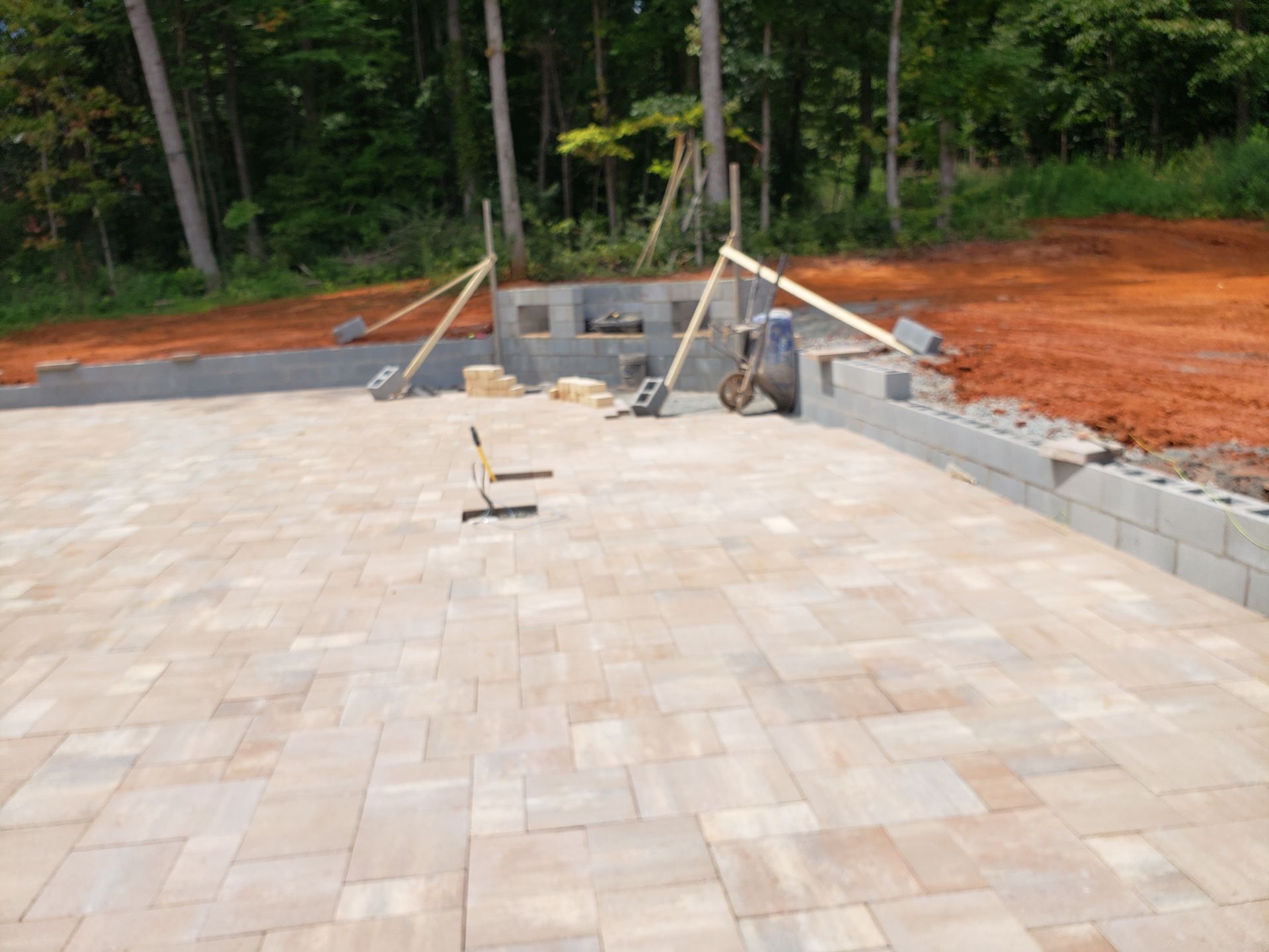 Patio under construction with pavers, retaining wall, and fireplace structure against a wooded backdrop.