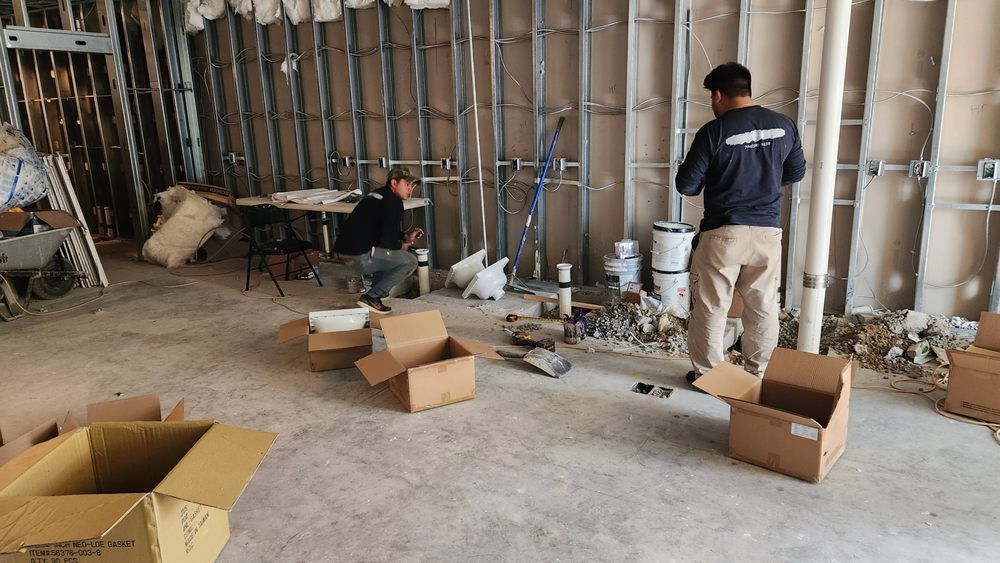 Two men working in a construction site with exposed framing, pipes, and cardboard boxes.