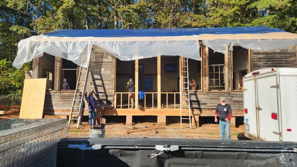 People renovating a weathered, wooden house. Blue tarp covers the roof. A truck bed is in the foreground.