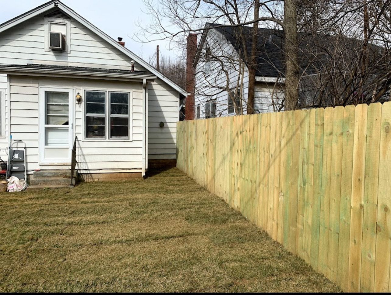 A wooden fence borders a lawn next to a white house with a small front porch and windows.