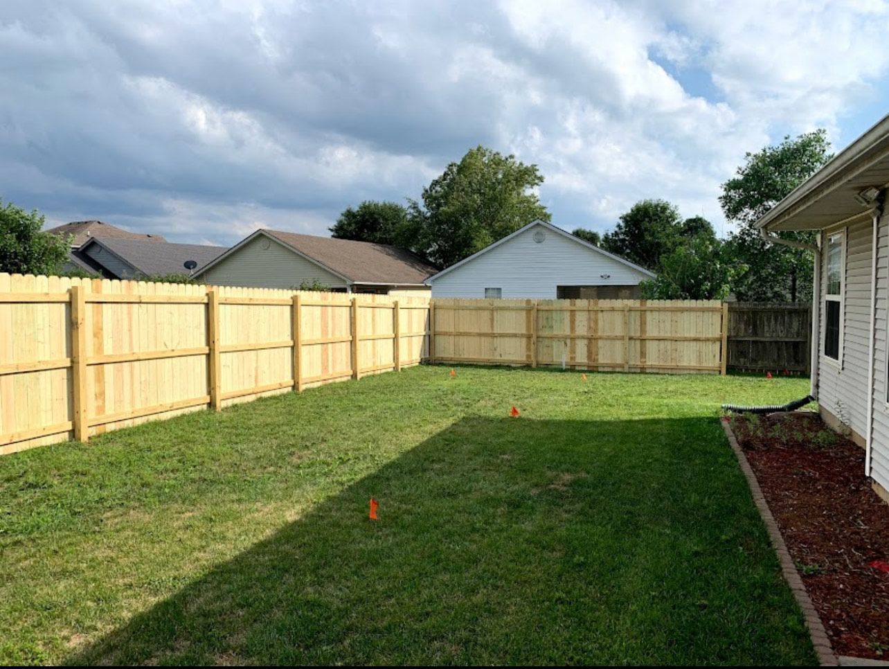 A backyard with a wooden fence, green grass, and houses in the background under a cloudy sky.