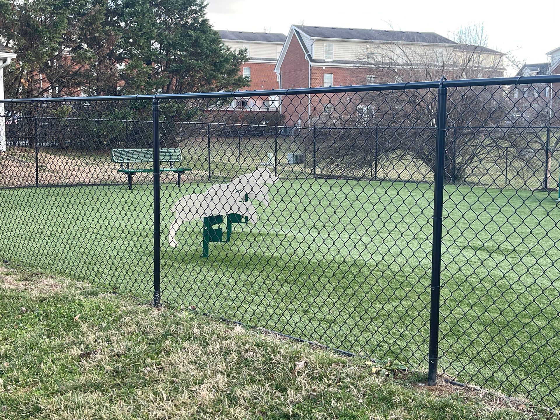 Black chain-link fence encloses a grassy dog park with a green waste bag dispenser and bench.
