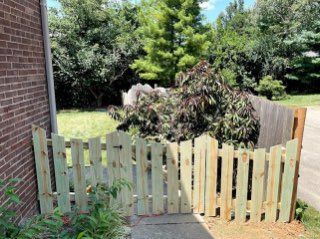 Wooden picket fence with an opening, next to a brick wall and foliage.