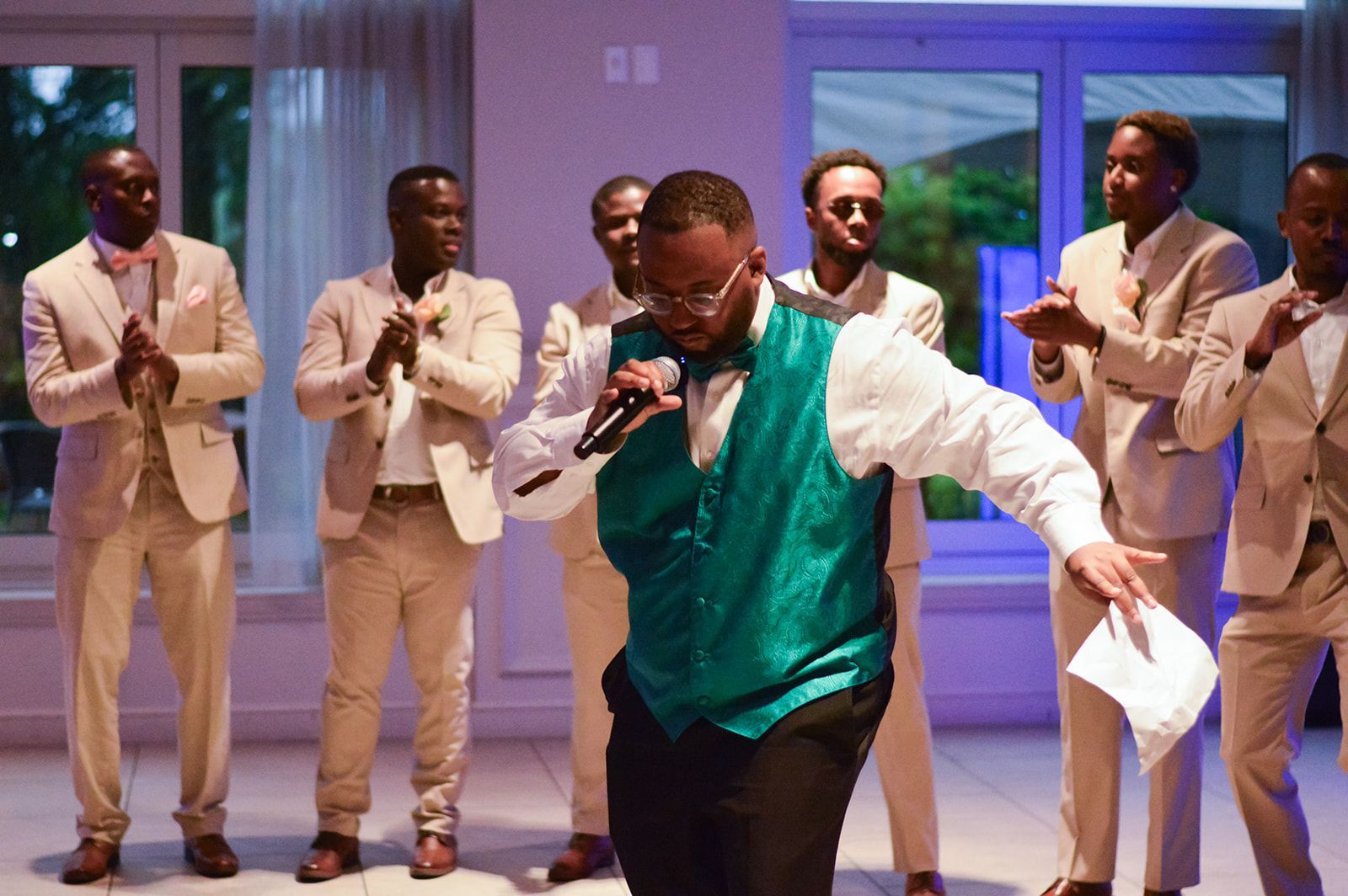 Un homme vêtu d'un gilet vert chante lors d'un événement, tenant un micro et du papier, entouré de six hommes en costumes beiges qui applaudissent.