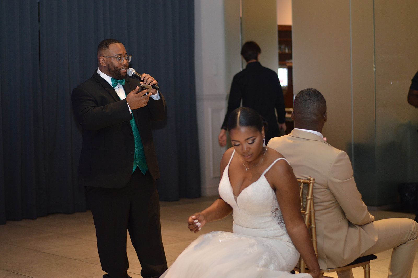 Un homme en smoking avec un micro prend la parole lors d'une réception de mariage. Les mariés jouent à un jeu assis.