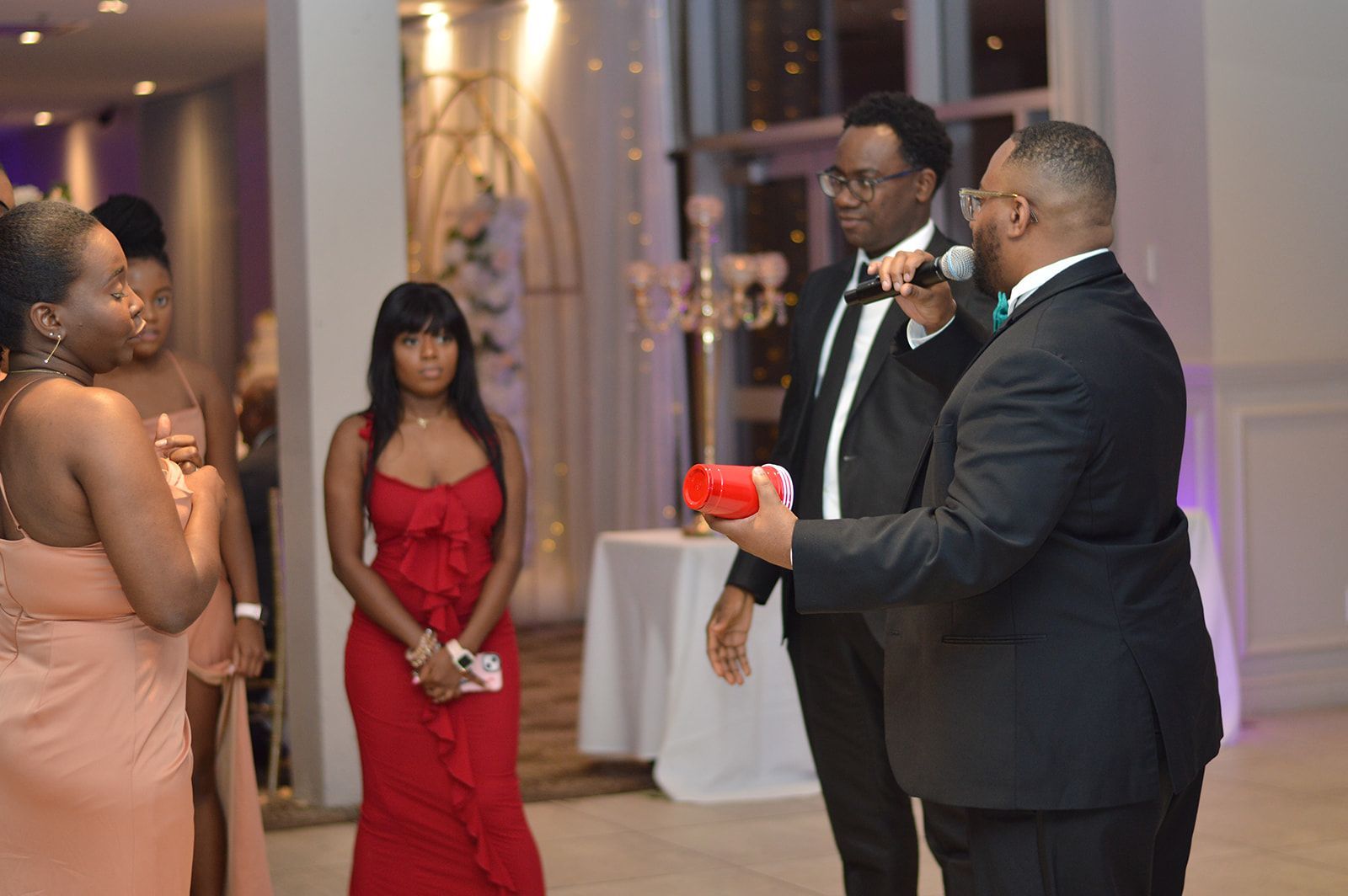 Des personnes en tenue formelle lors d'un événement, un homme parle dans un microphone, tenant un objet rouge, tandis que d'autres écoutent.