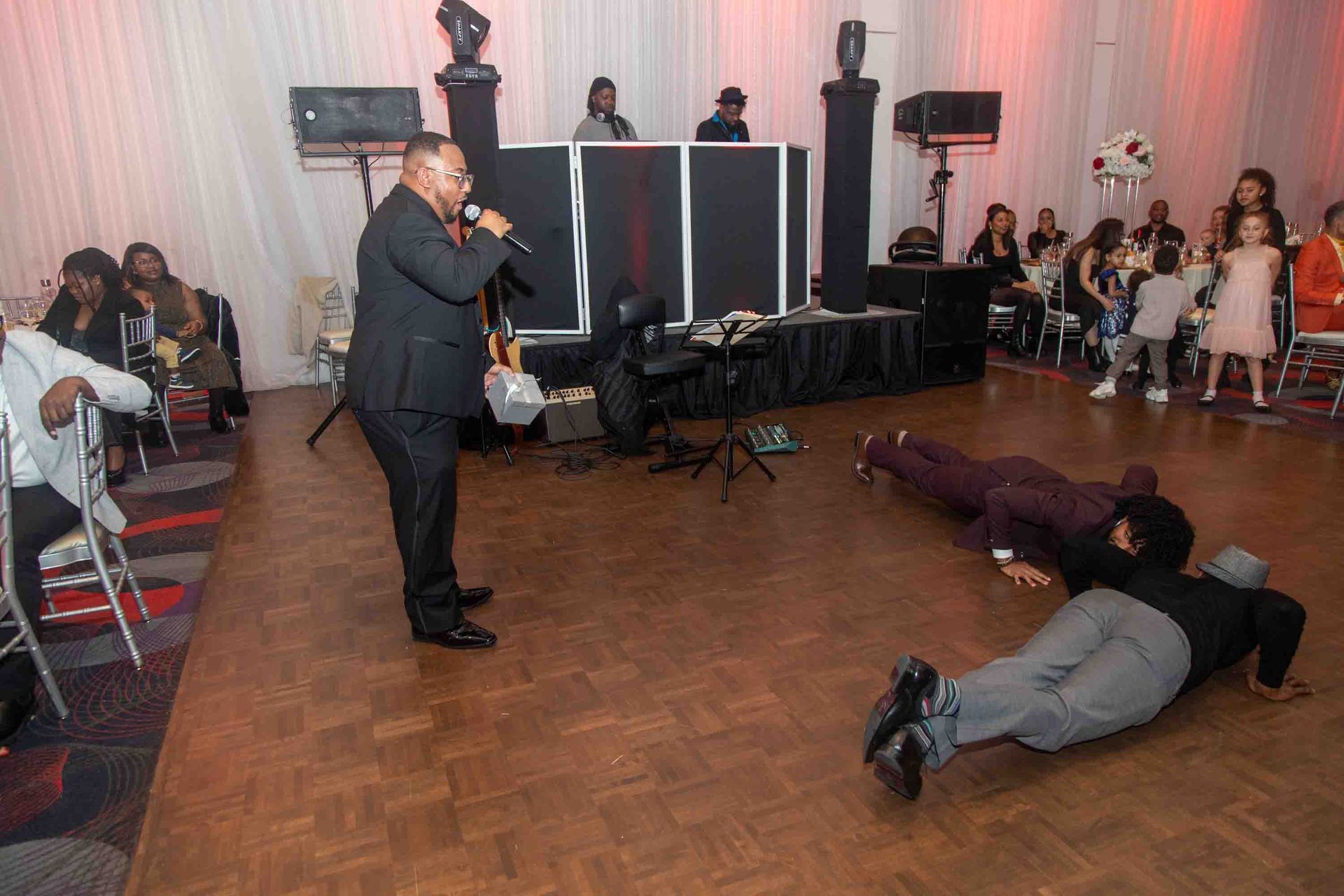 Un homme chante au micro lors d'une fête tandis que deux hommes font des pompes sur la piste de danse. Les gens regardent.