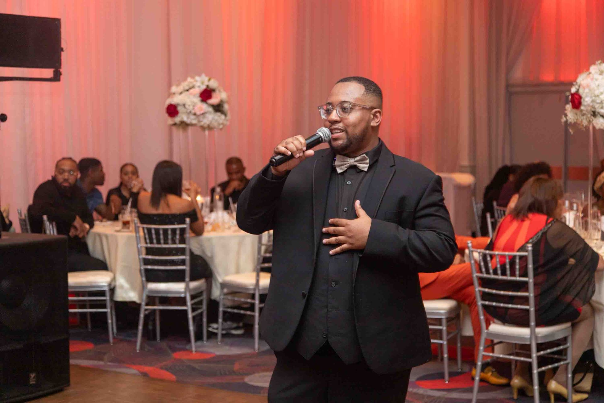 Un homme noir en costume chante au micro lors d'un événement. Il se tient devant des tables rondes avec des invités.