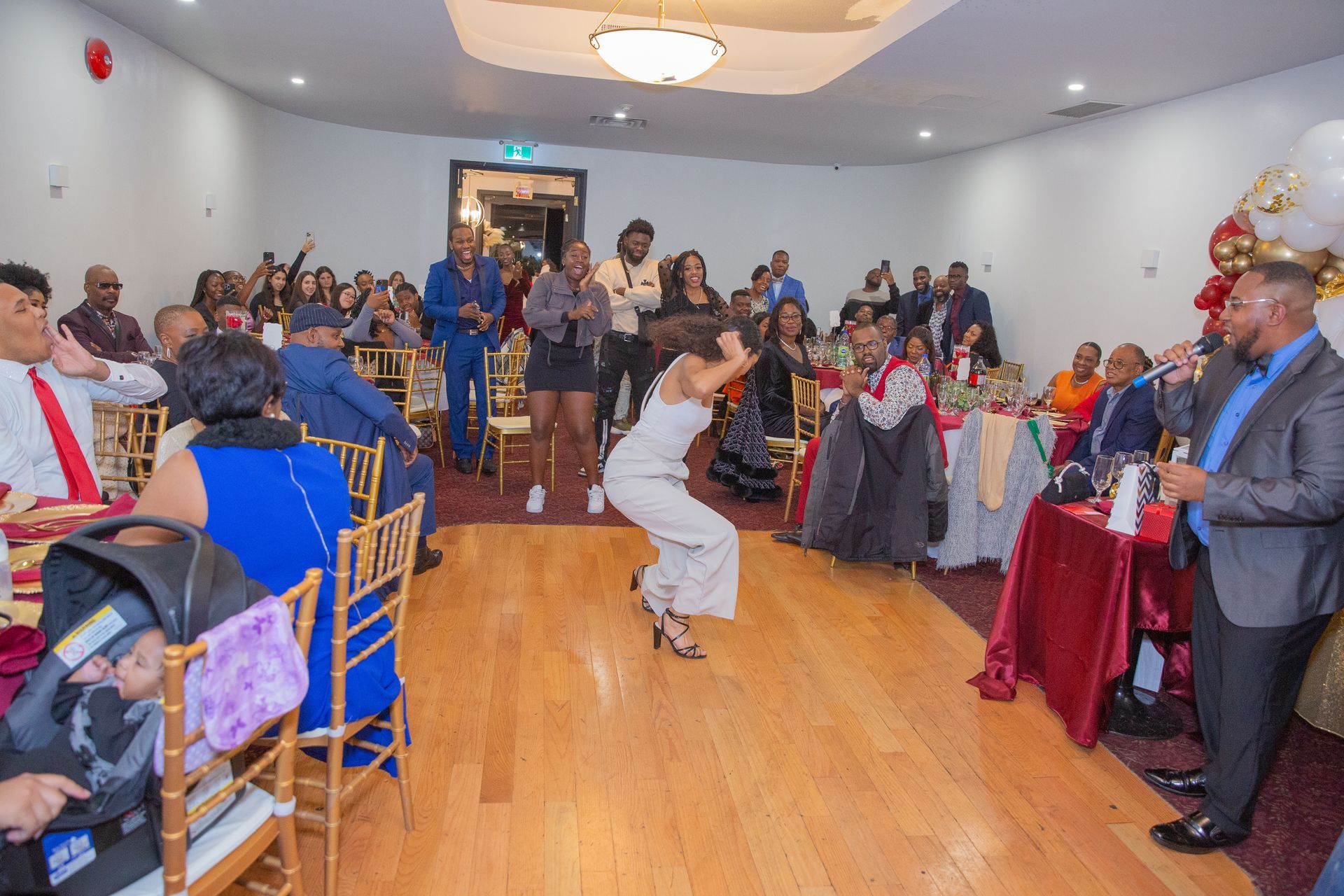 Un événement animé avec une femme dansant sur un plancher de bois. Les gens sont rassemblés autour de tables dans une salle lumineuse, en fête.