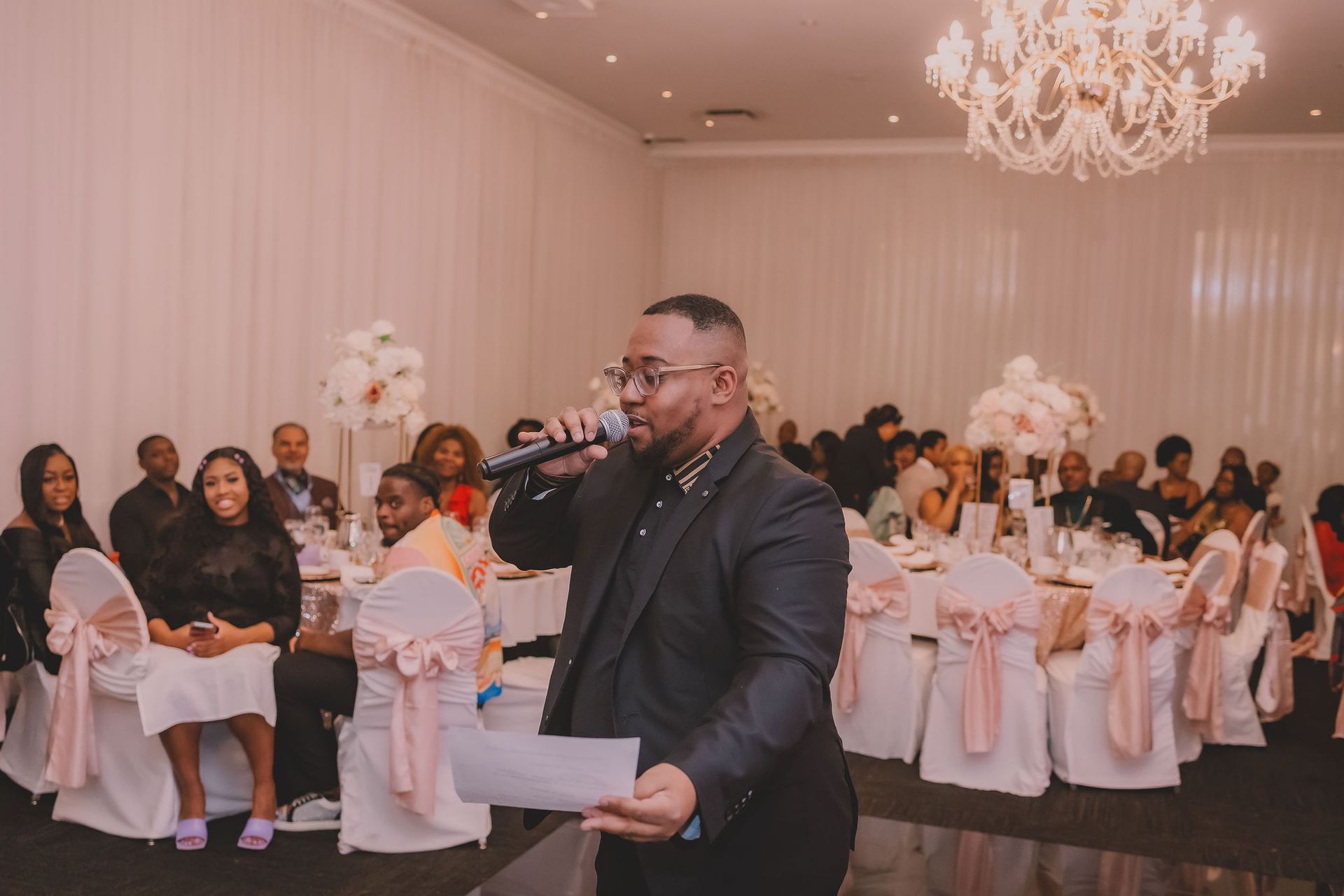 Lors d'un événement officiel, un homme en costume noir s'adresse à des invités assis au micro. Les tables sont décorées de rose et un lustre est suspendu au-dessus.