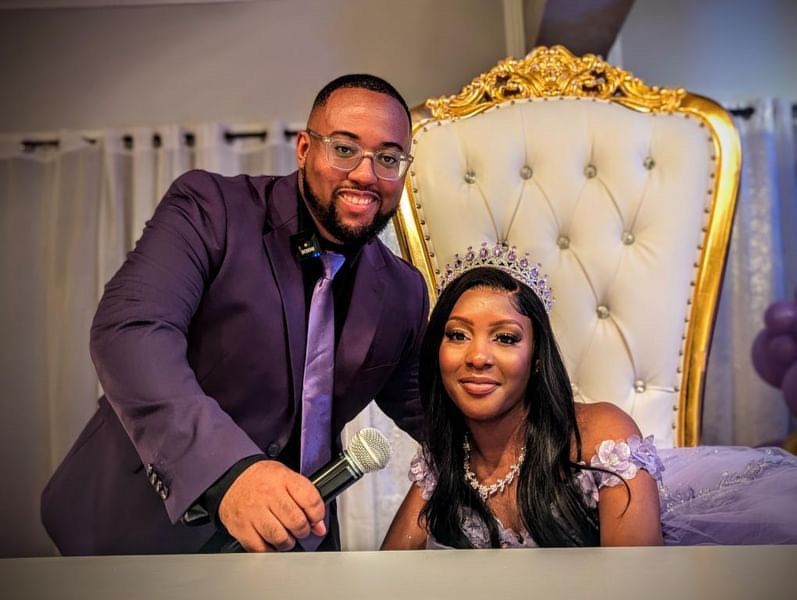 Un homme en costume violet tient un microphone, sourit à côté d'une femme portant un diadème et une robe violette, assise sur un trône en or.
