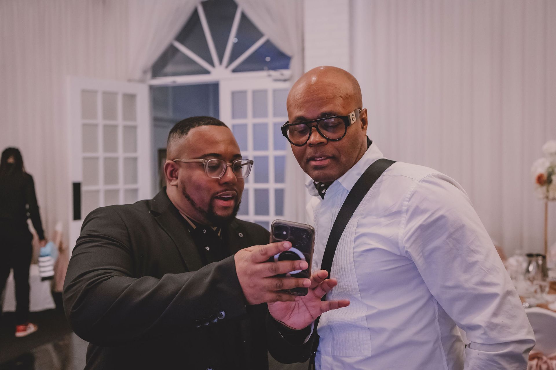 Deux hommes regardant un téléphone, l'un en blazer, l'autre en chemise blanche, à l'intérieur près des portes-fenêtres.