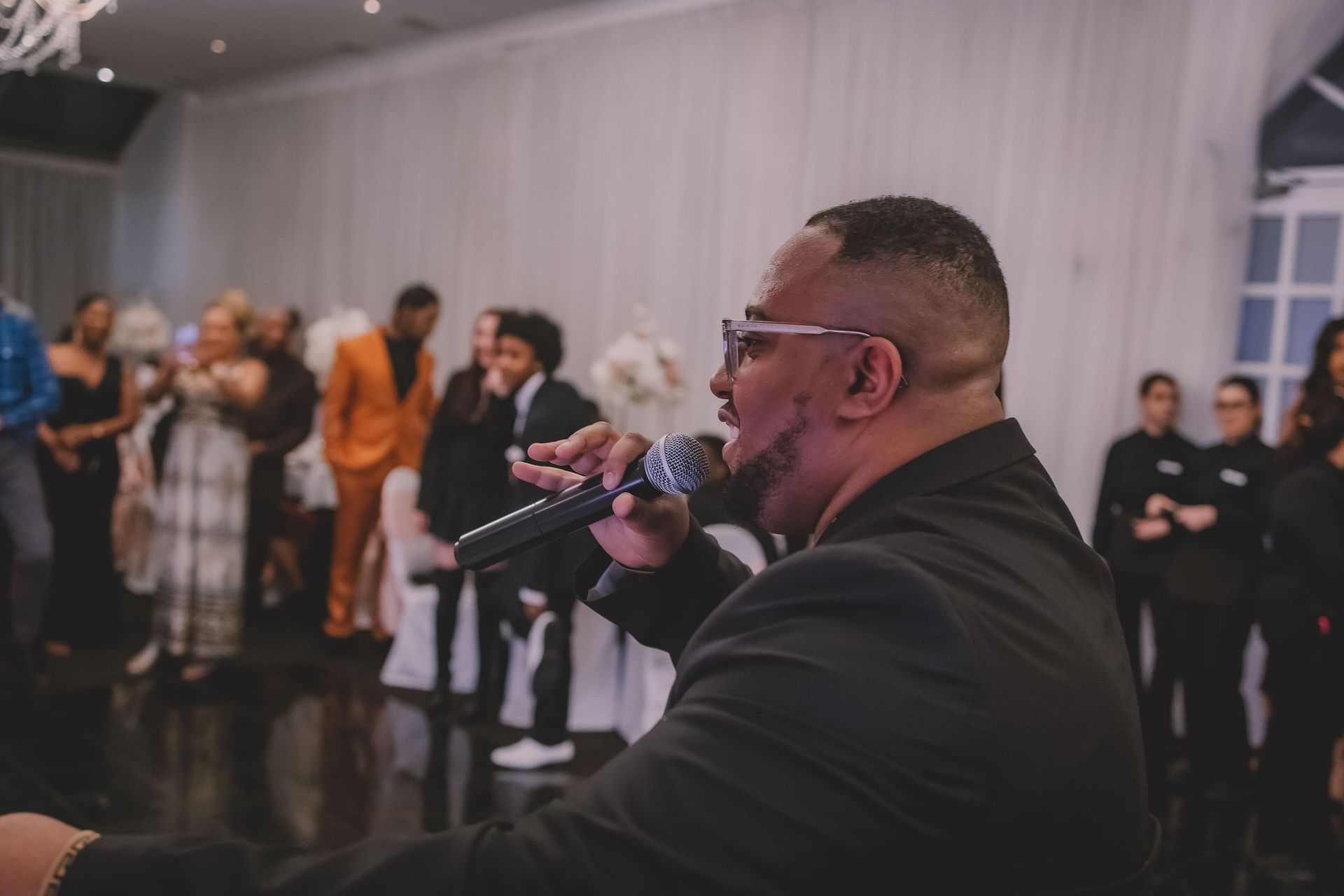 Un homme en costume parle au micro lors d'un événement, avec des invités en arrière-plan. Il porte des lunettes et affiche une expression sérieuse.