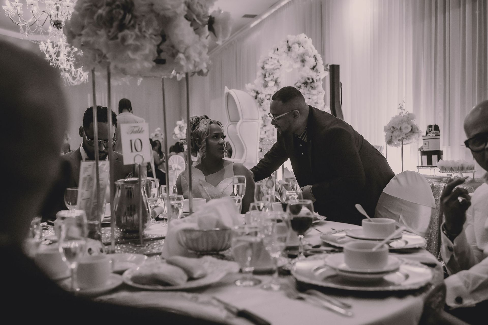 Un homme en costume se penche vers une femme à une table de réception de mariage. On aperçoit des compositions florales blanches et des tables dressées avec de la porcelaine.