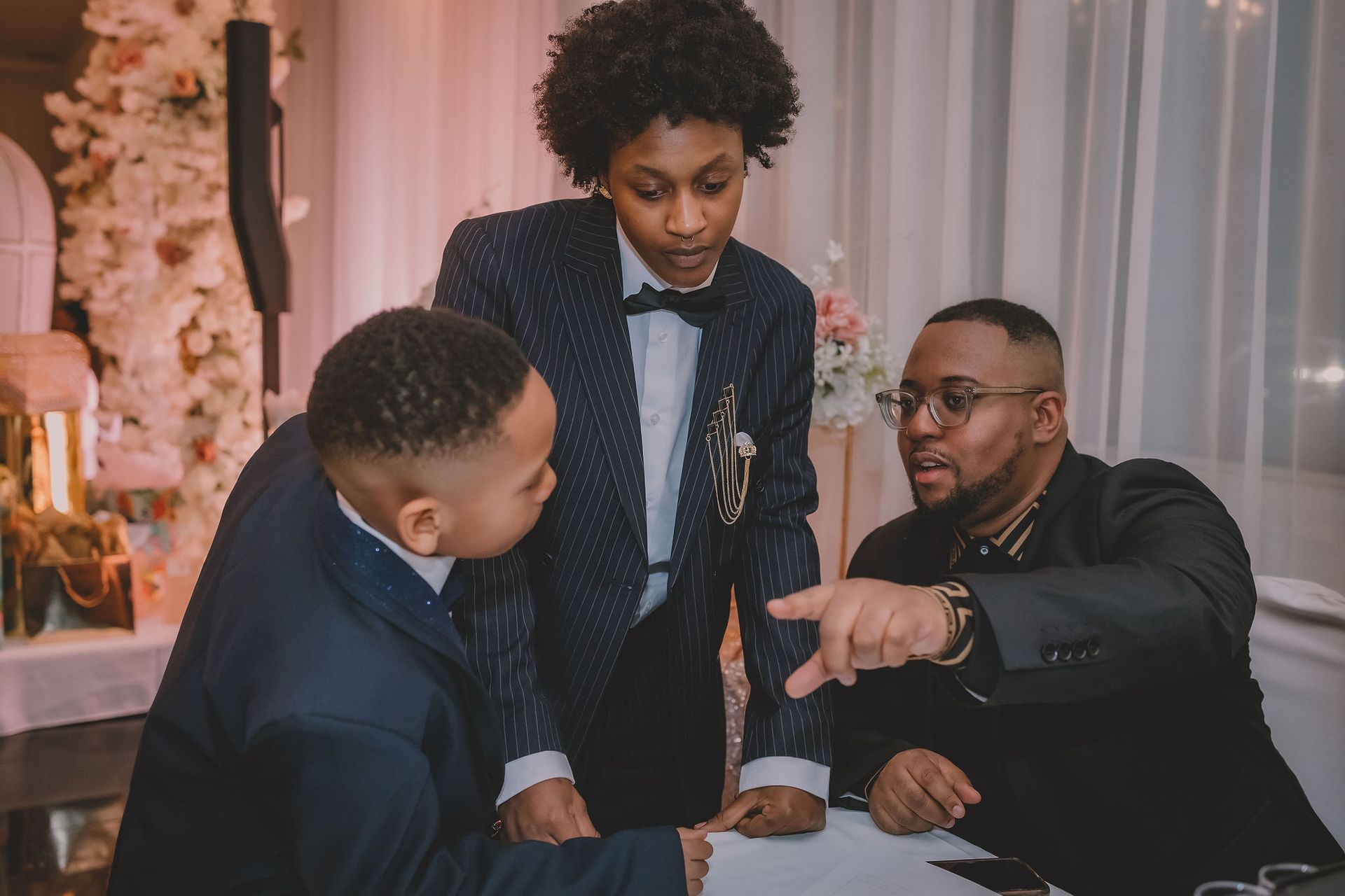 Trois hommes en tenue formelle discutent à une table ; l'un d'eux montre du doigt. Intérieur élégant avec décorations florales.