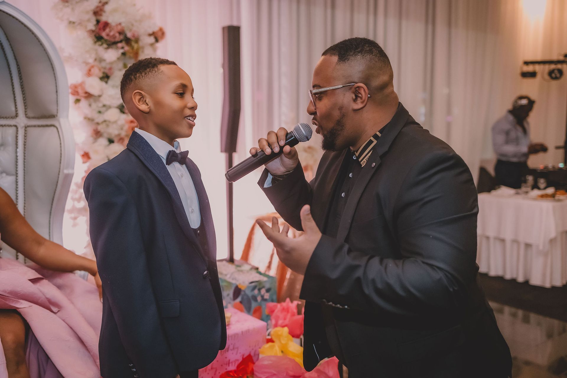 Un homme parle dans un microphone, face à un jeune garçon en costume et nœud papillon ; lors d'une fête.