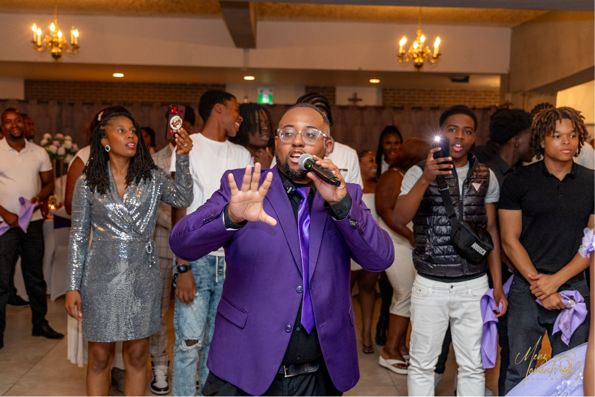 Un homme en costume violet chantant dans un microphone, entouré d'une foule de personnes dans une pièce bien éclairée.