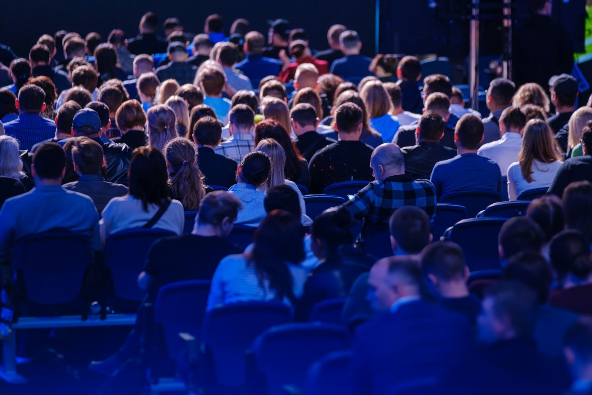 Public assis dans un grand auditorium, vu de dos, avec de nombreuses personnes présentes. Éclairage bleu et focus sur la scène.